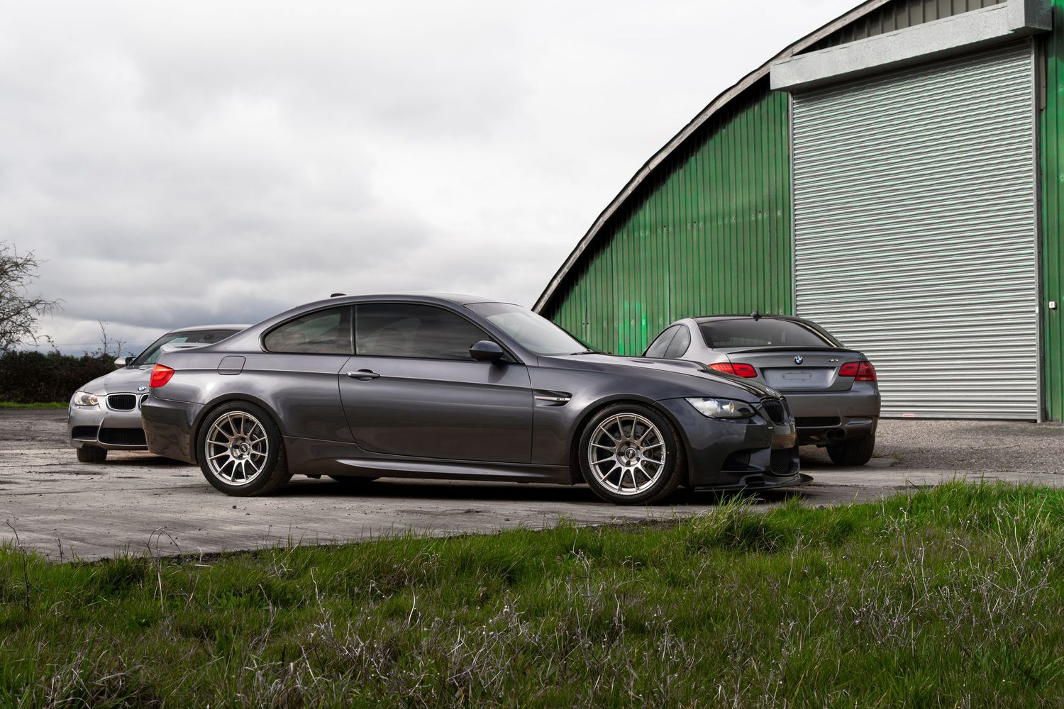 Two gray BMW M3 coupes parked back to back outside a large green arched hangar.