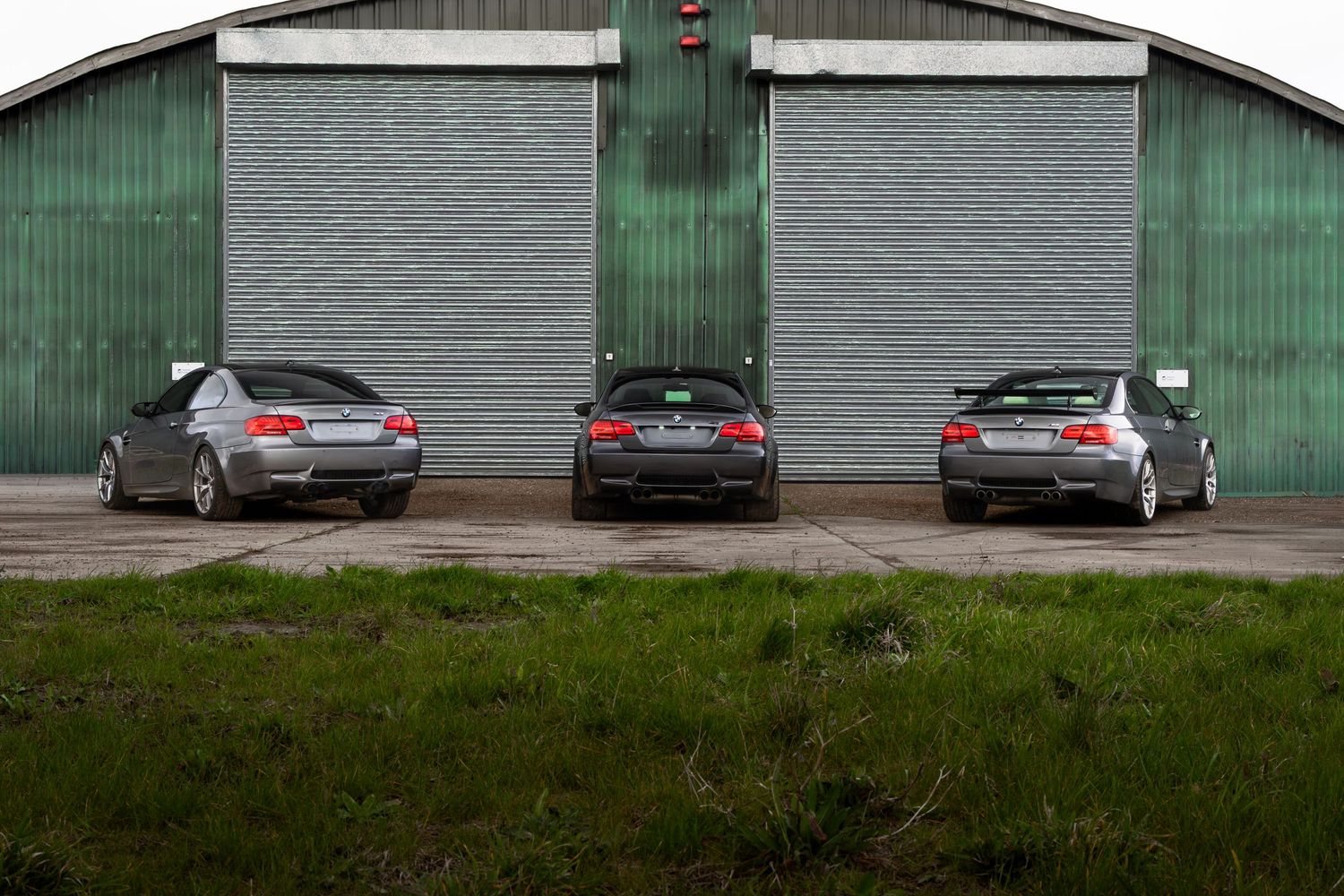 Three BMW M3 coupes parked rear-facing in front of green corrugated metal garage doors.