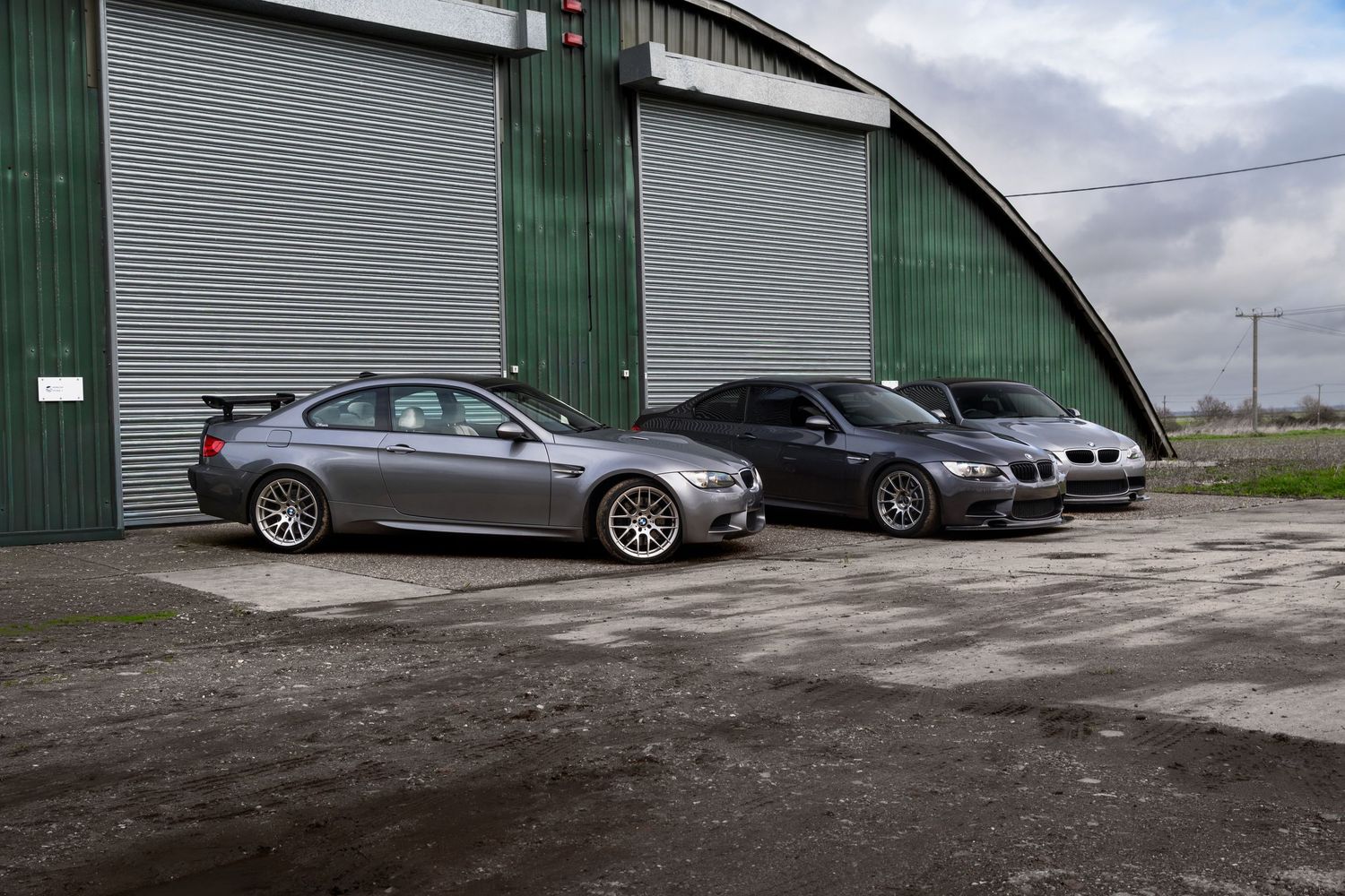 Two grey BMW M3 coupes parked side by side outside a large green hangar.