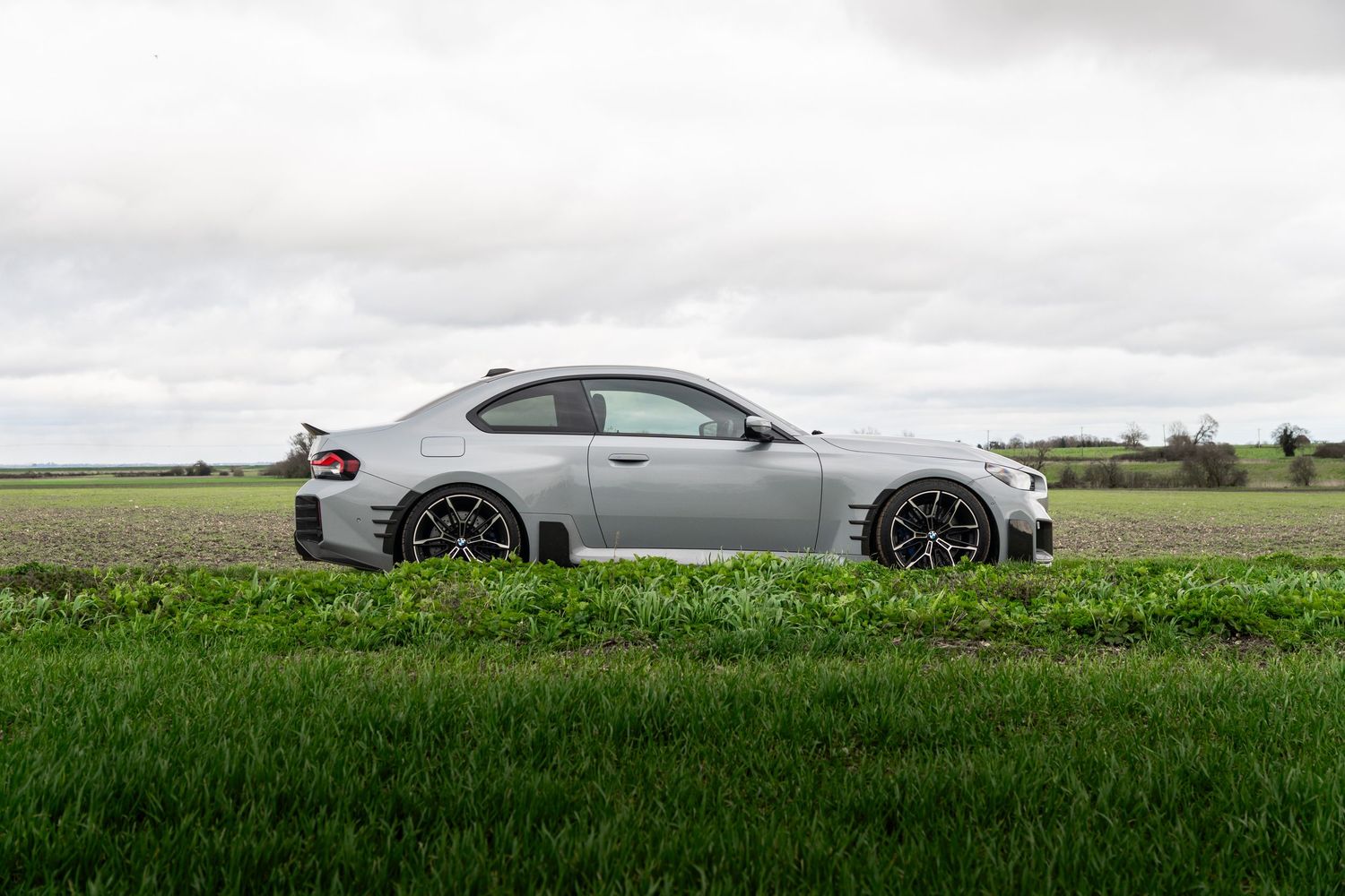 Matte gray BMW M2 Competition coupe posed on a rural road with green fields in background.