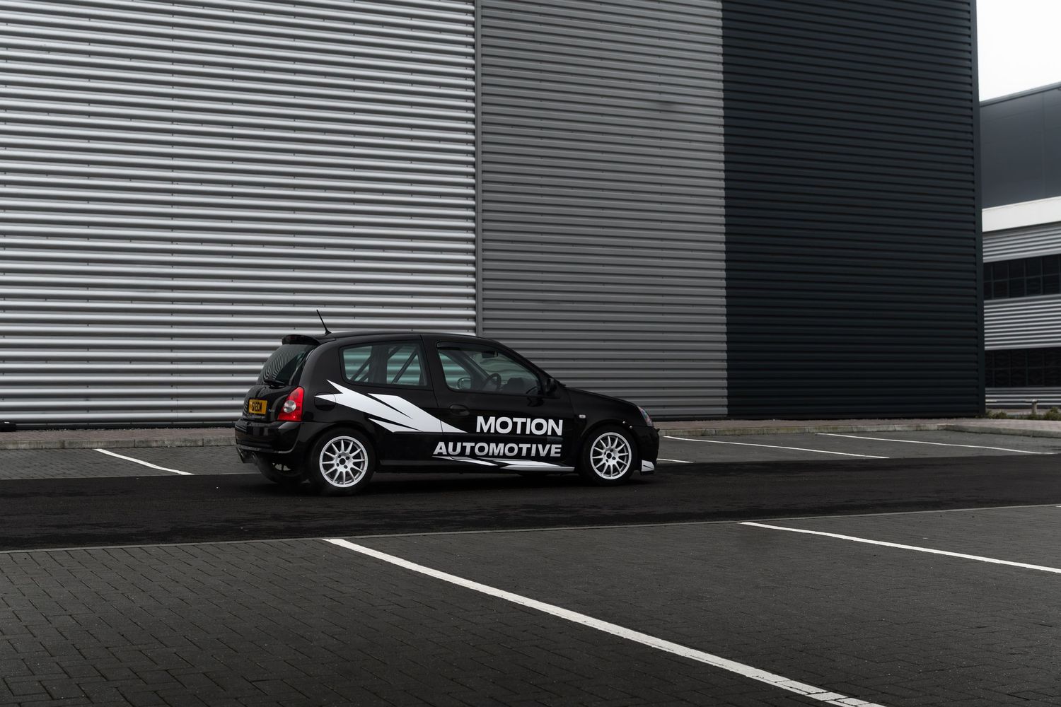 Rear view of black Motion Automotive wrapped Renault Clio parked in lot by industrial building.