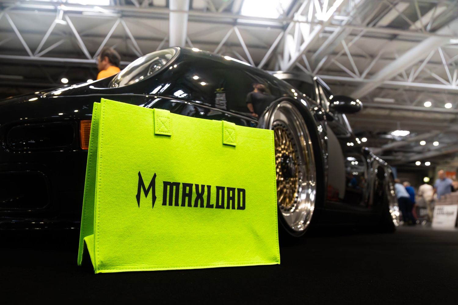 Neon green Maxload shopping bag displayed in front of a black modified car at an auto show.