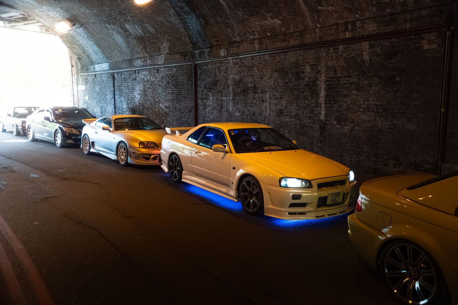 Row of modified cars including yellow Nissan Skyline GT-R lined up in tunnel.