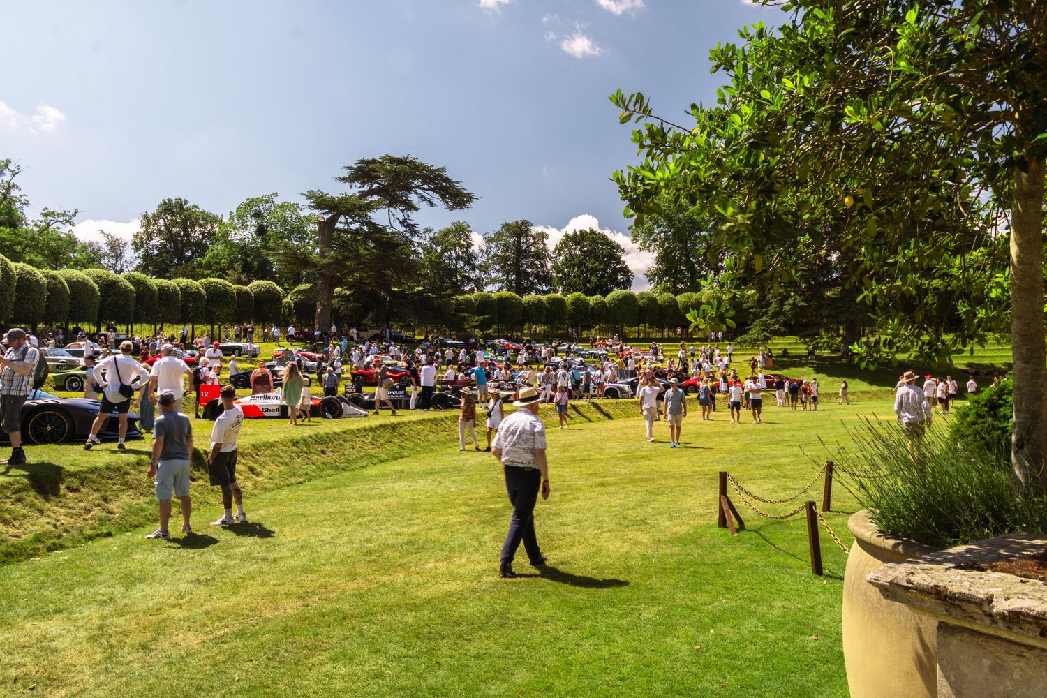 Crowds gather on lush green lawns at a sunny outdoor garden event surrounded by trees and hedges.