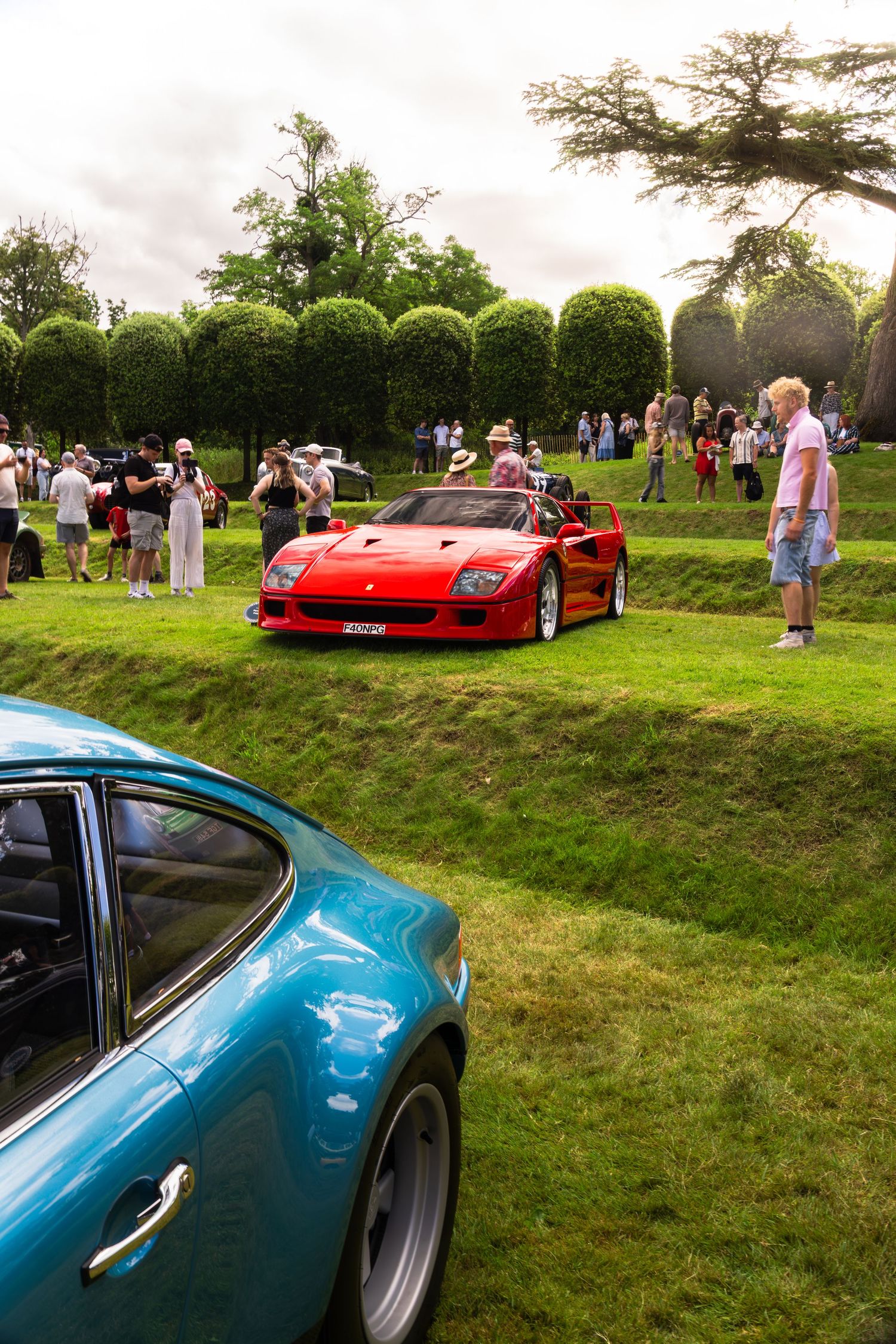 Red Ferrari F40 supercar on display at an outdoor classic car show, with spectators and manicured hedges in background.