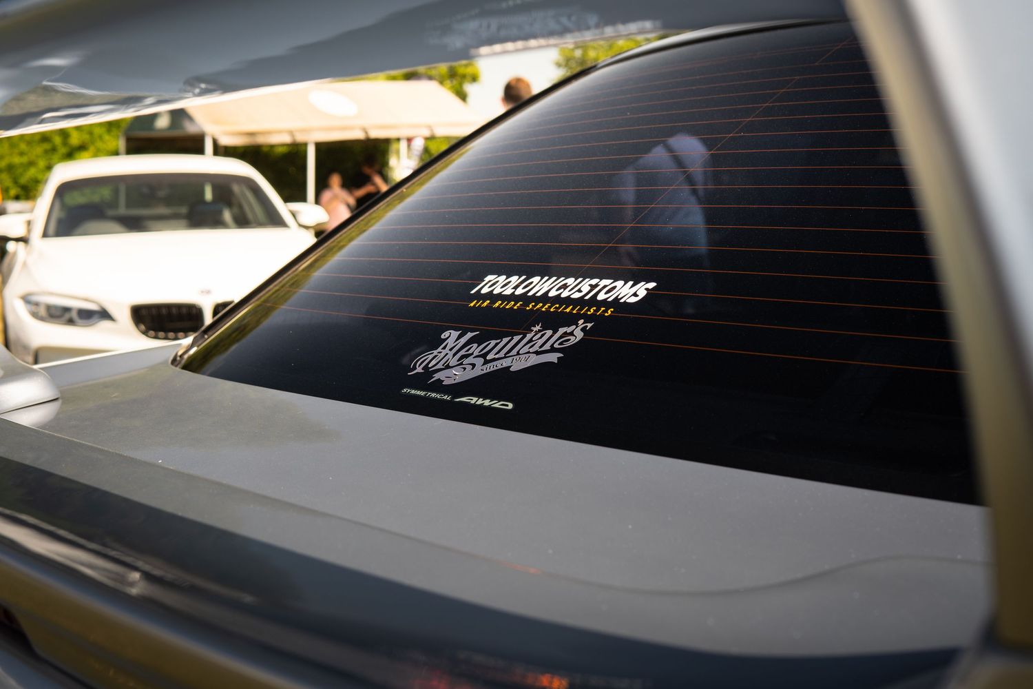 Gray car rear window with Dox Customs and Meguiars vinyl decals at automotive meet.