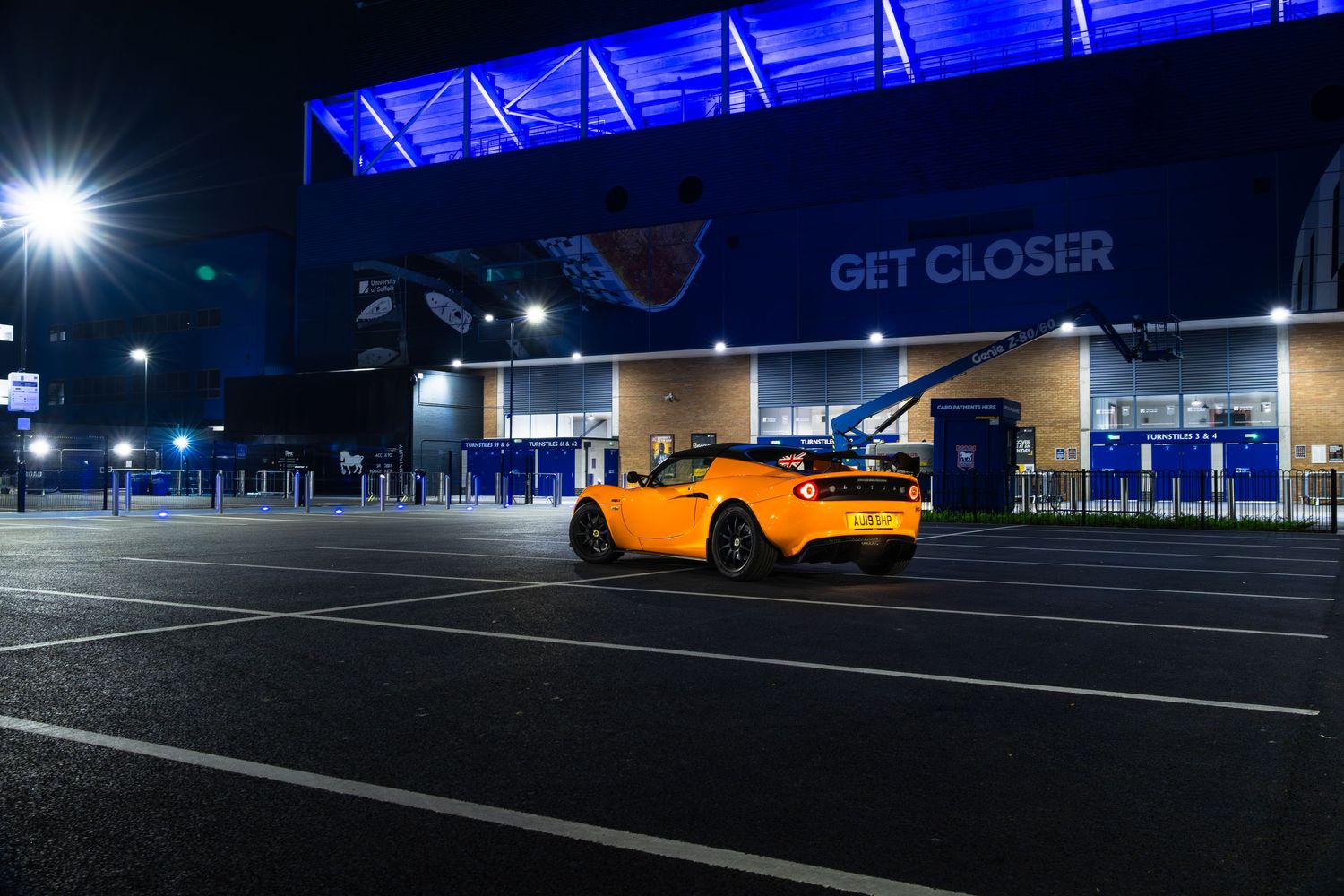 Orange Lotus Elise roadster photographed at night beside brightly lit blue football stadium exterior.