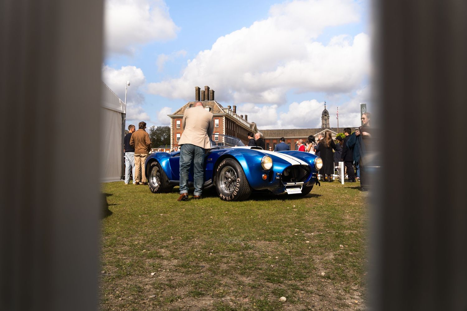Blue Shelby Cobra replica car viewed through a gap at an outdoor automotive show.