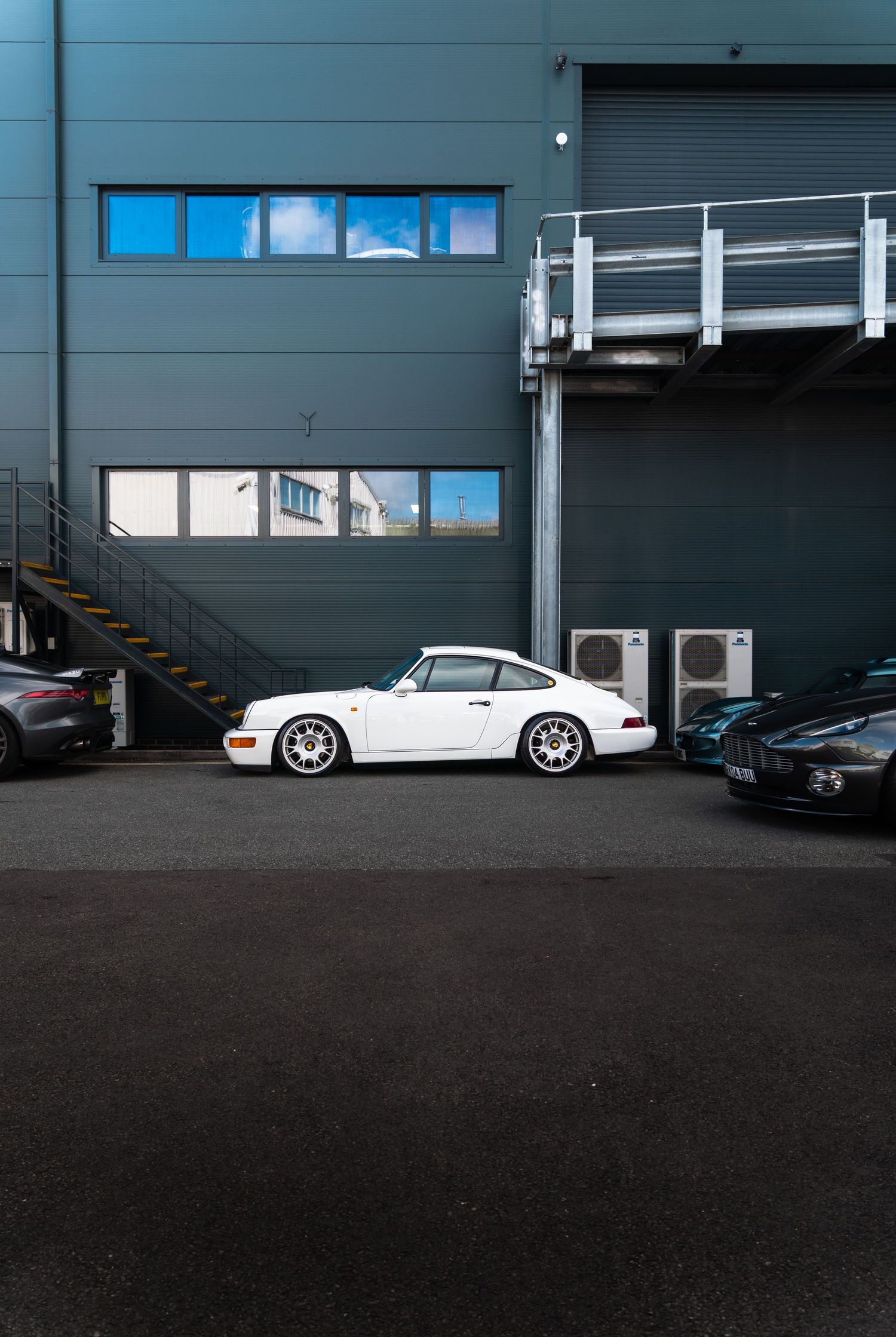 Classic white Porsche 911 parked outside a grey industrial building with metal stairs and walkway.