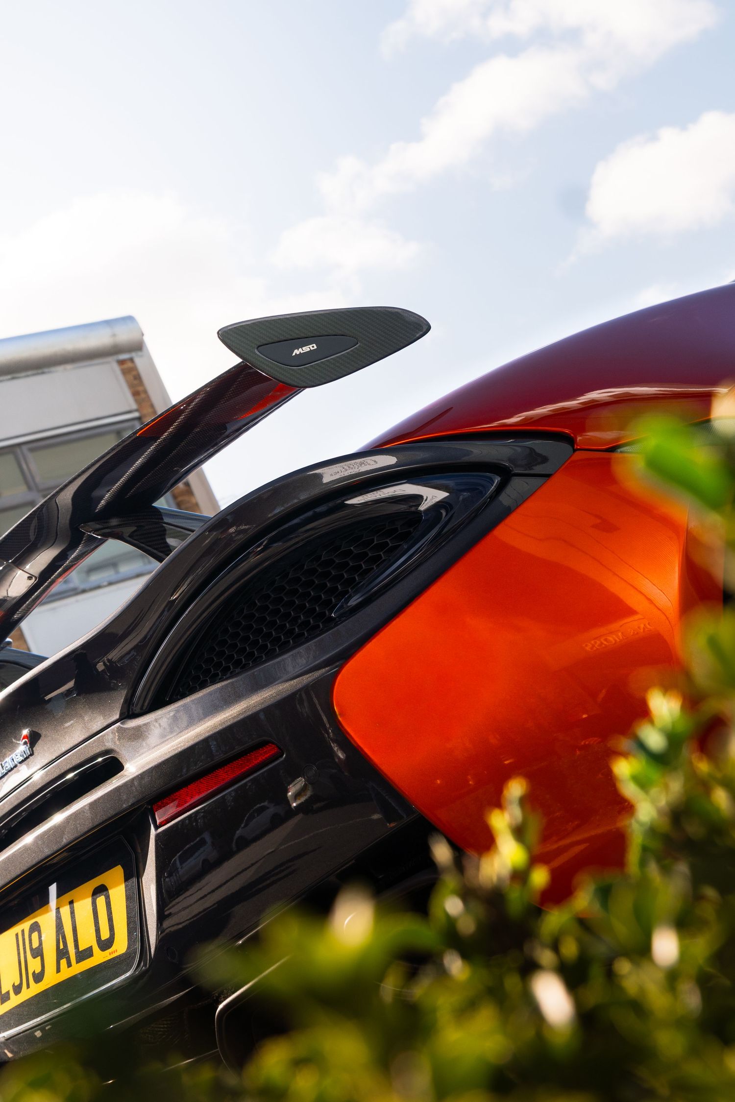 Close-up rear view of a vibrant orange McLaren supercar with UK license plate parked near green hedges.