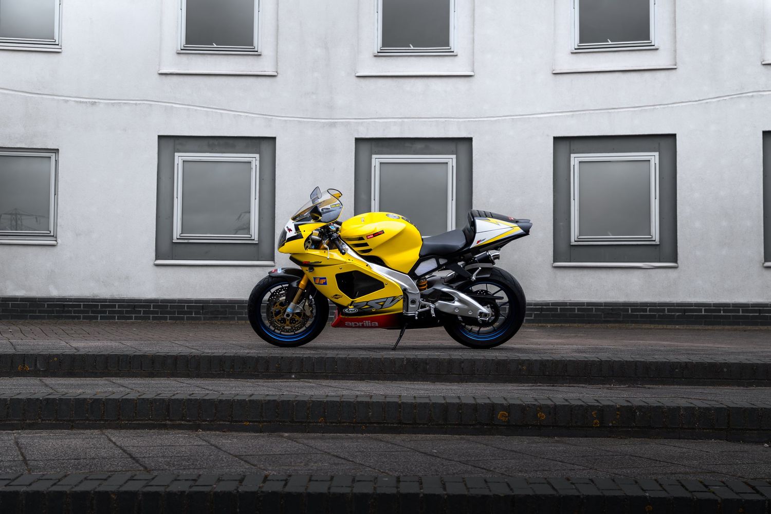 Yellow Aprilia RSV-R superbike parked on brick pavement beside a white building with grey-framed windows.