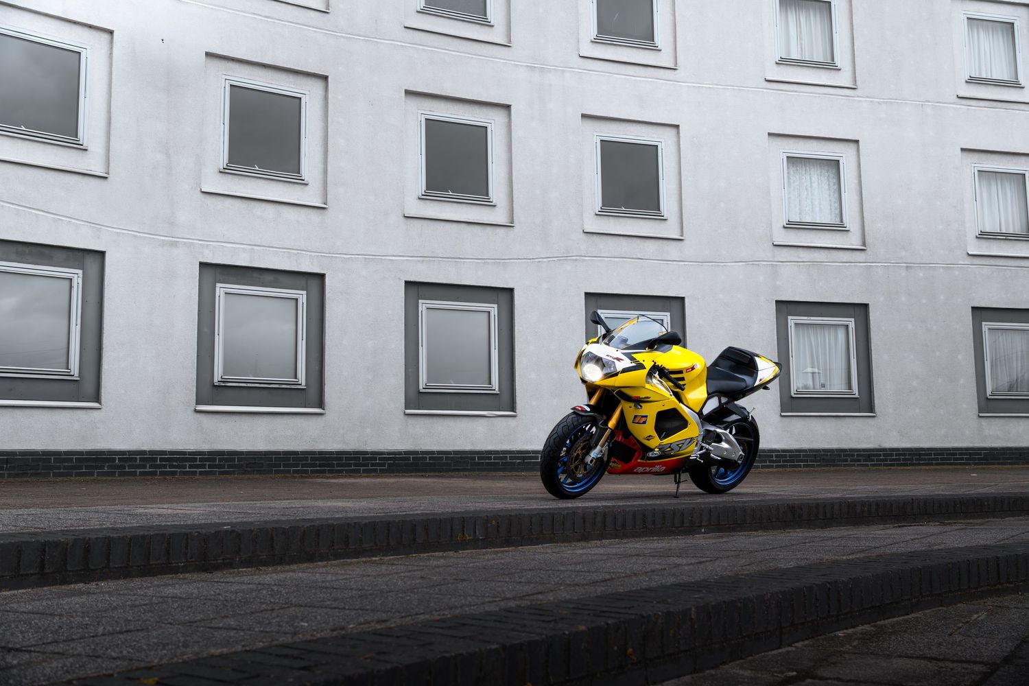 Yellow Aprilia RSV1000 superbike parked on stepped pavement against a white multi-windowed building.