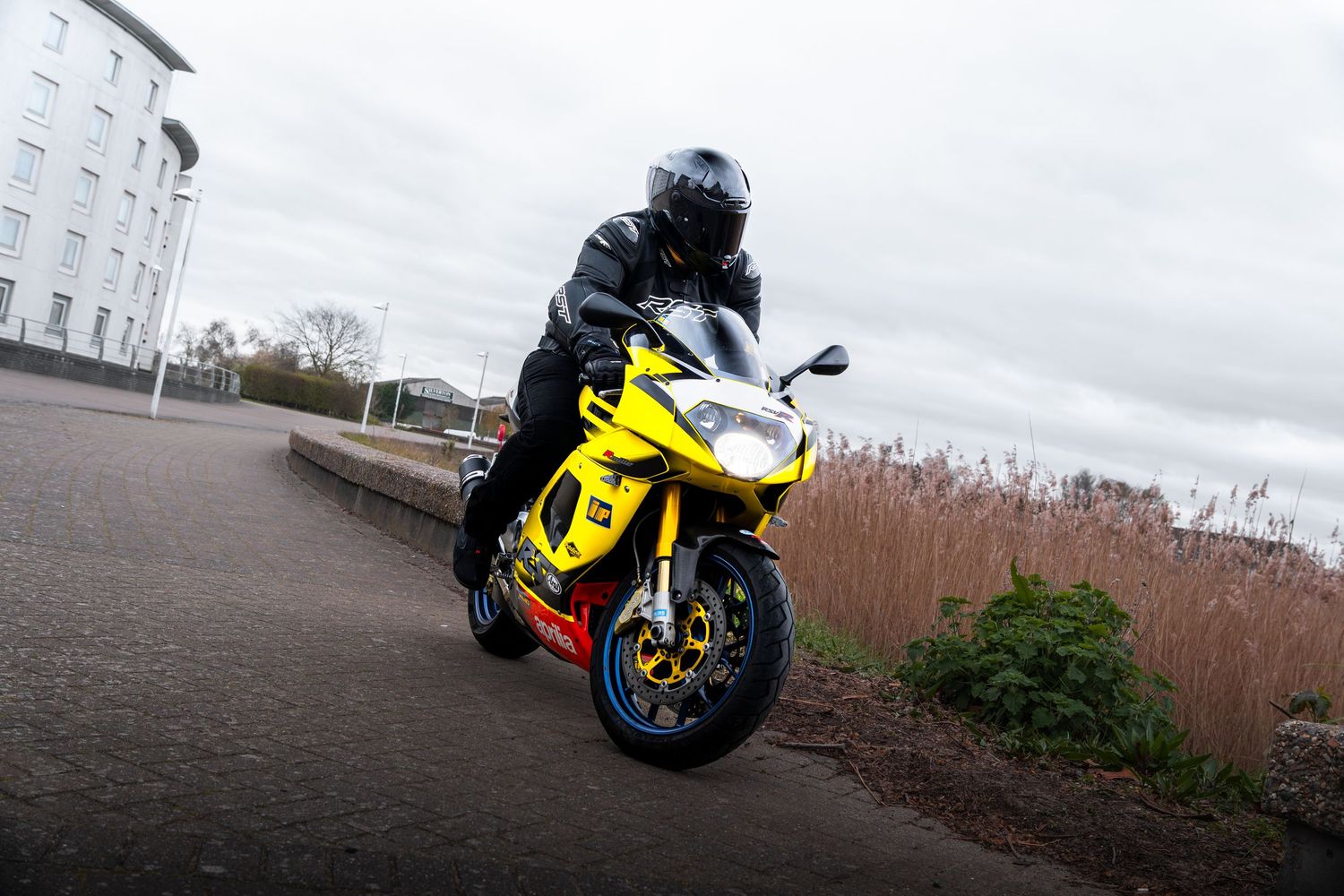 Yellow Aprilia RSV sportbike ridden by a black-suited motorcyclist on a road near water.