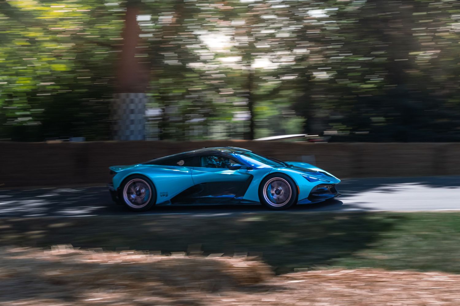A vibrant blue Aston Martin supercar speeds along a tree-lined road, captured in motion blur at a motorsport event.