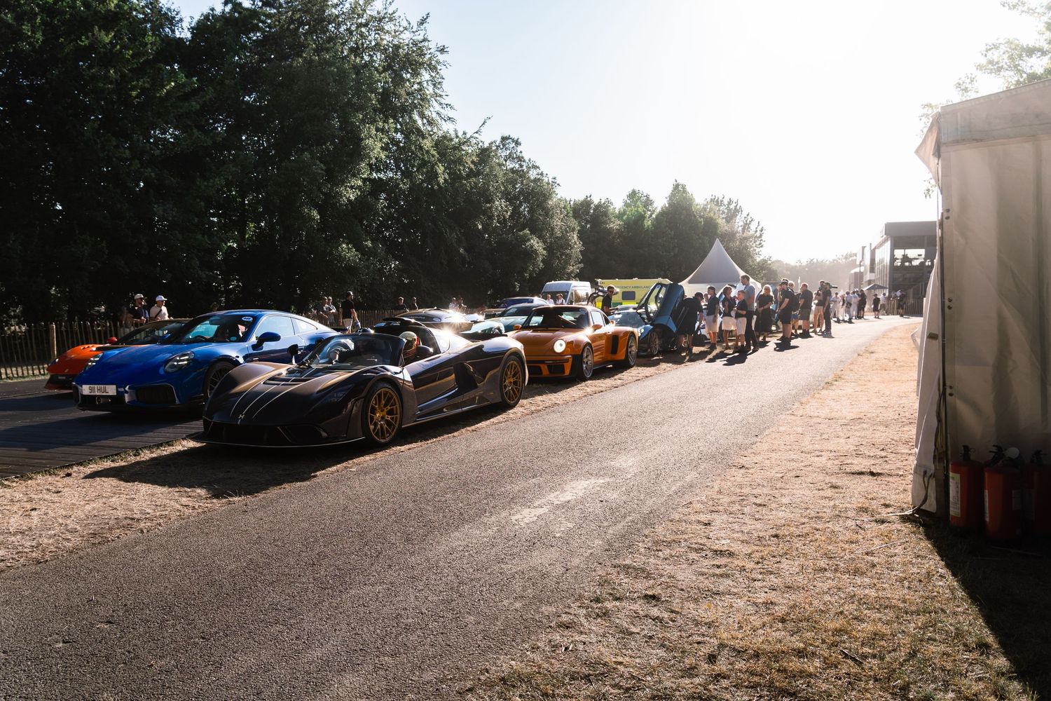 Luxury supercars lined up on a road at an outdoor motorsport event, surrounded by trees and spectators on a sunny day.