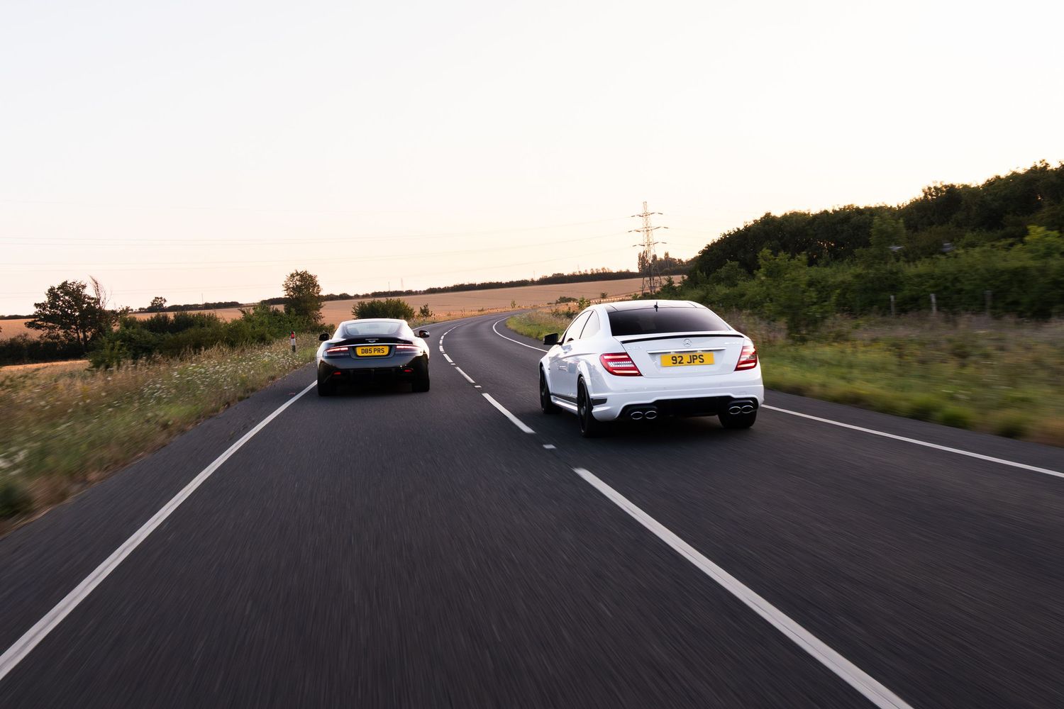 Two cars, one white and one black, driving on a rural road at dusk surrounded by green trees and golden fields.