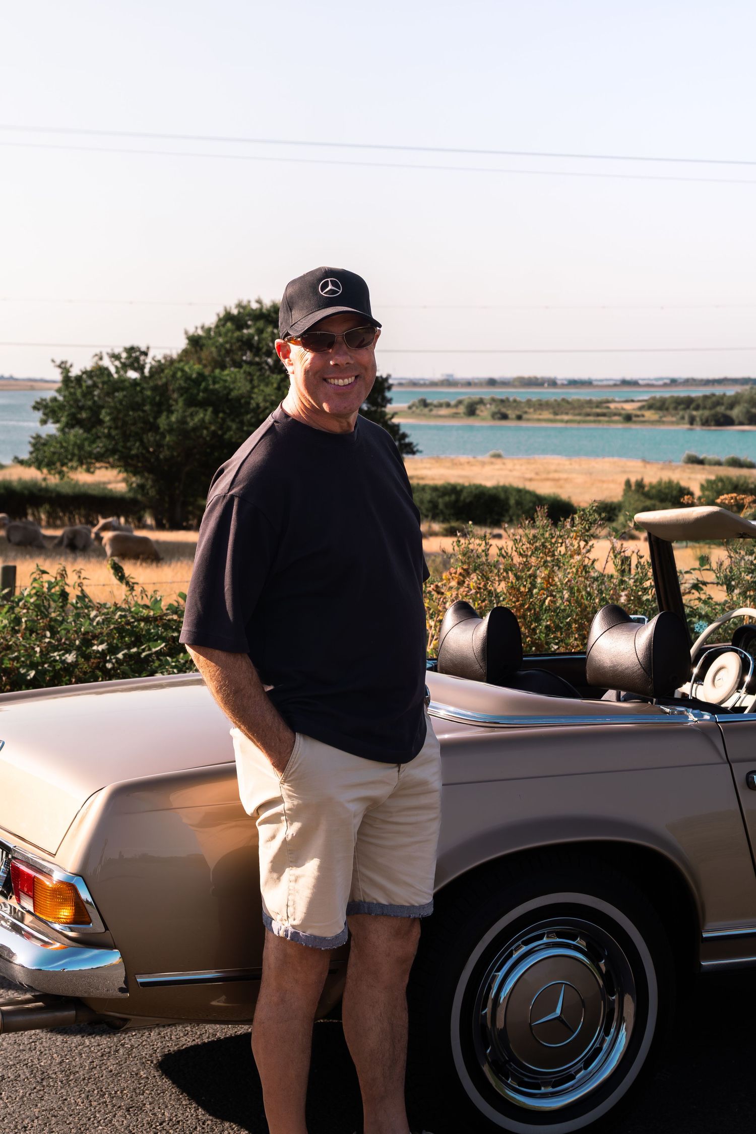 Man in navy Mercedes cap and shorts posing beside a vintage Mercedes-Benz convertible near a scenic waterfront.