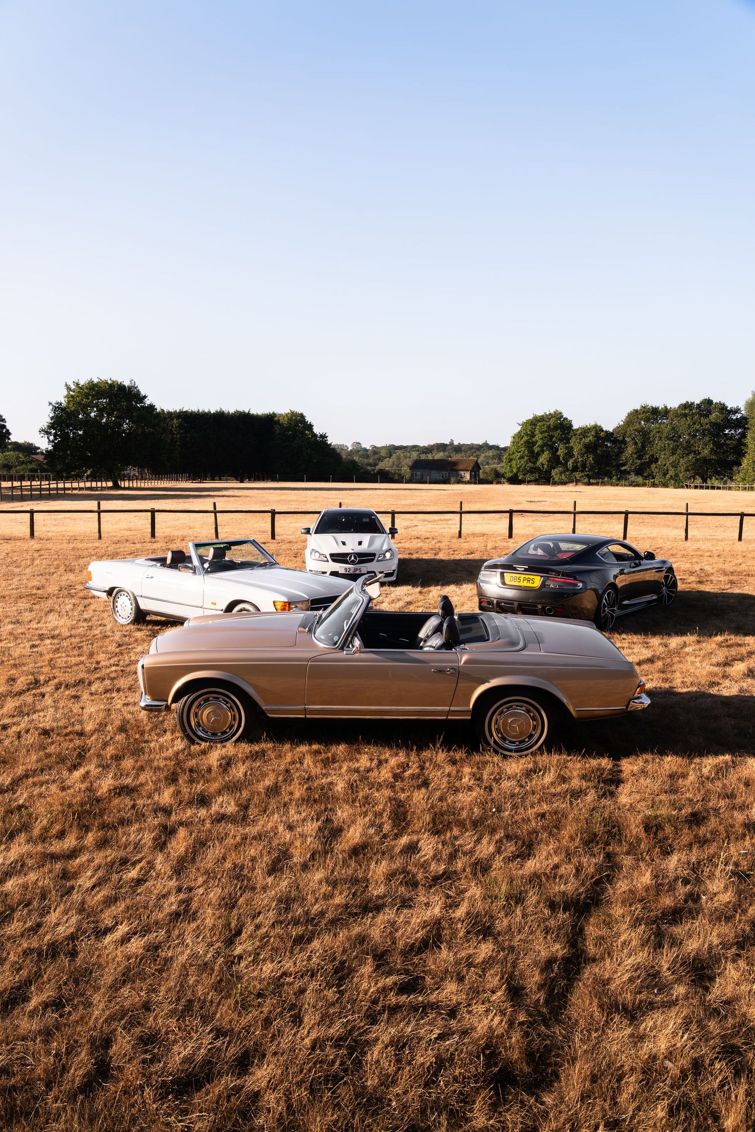 Three classic and modern convertible cars parked in a golden field with wooden fencing and trees under a clear blue sky.