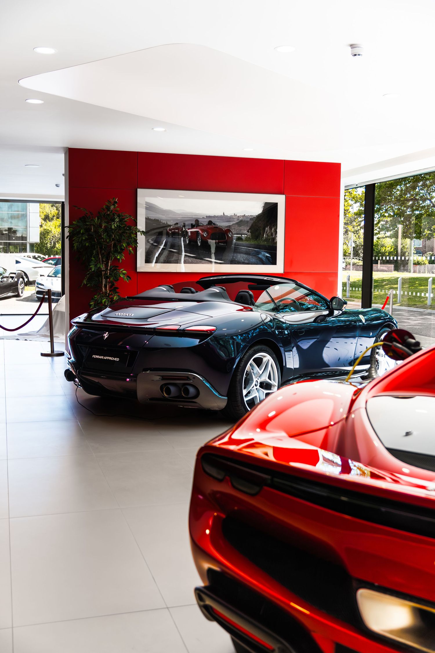 Ferrari dealership showroom featuring a dark green and red supercar on display with a racing artwork on the red accent wall.