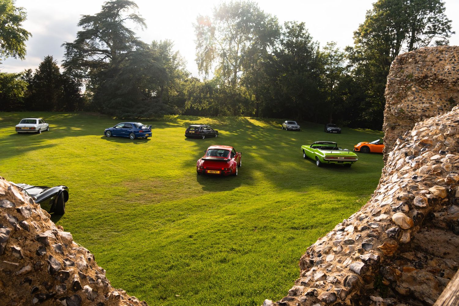 Classic cars parked on lush green grass surrounded by ancient stone ruins and tall trees at a vintage car show.