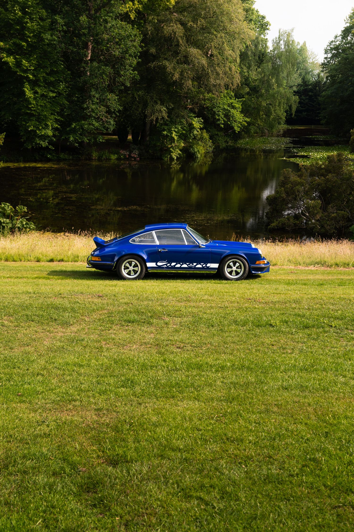A classic blue Porsche 911 Carrera parked on lush green grass beside a tranquil pond surrounded by trees.