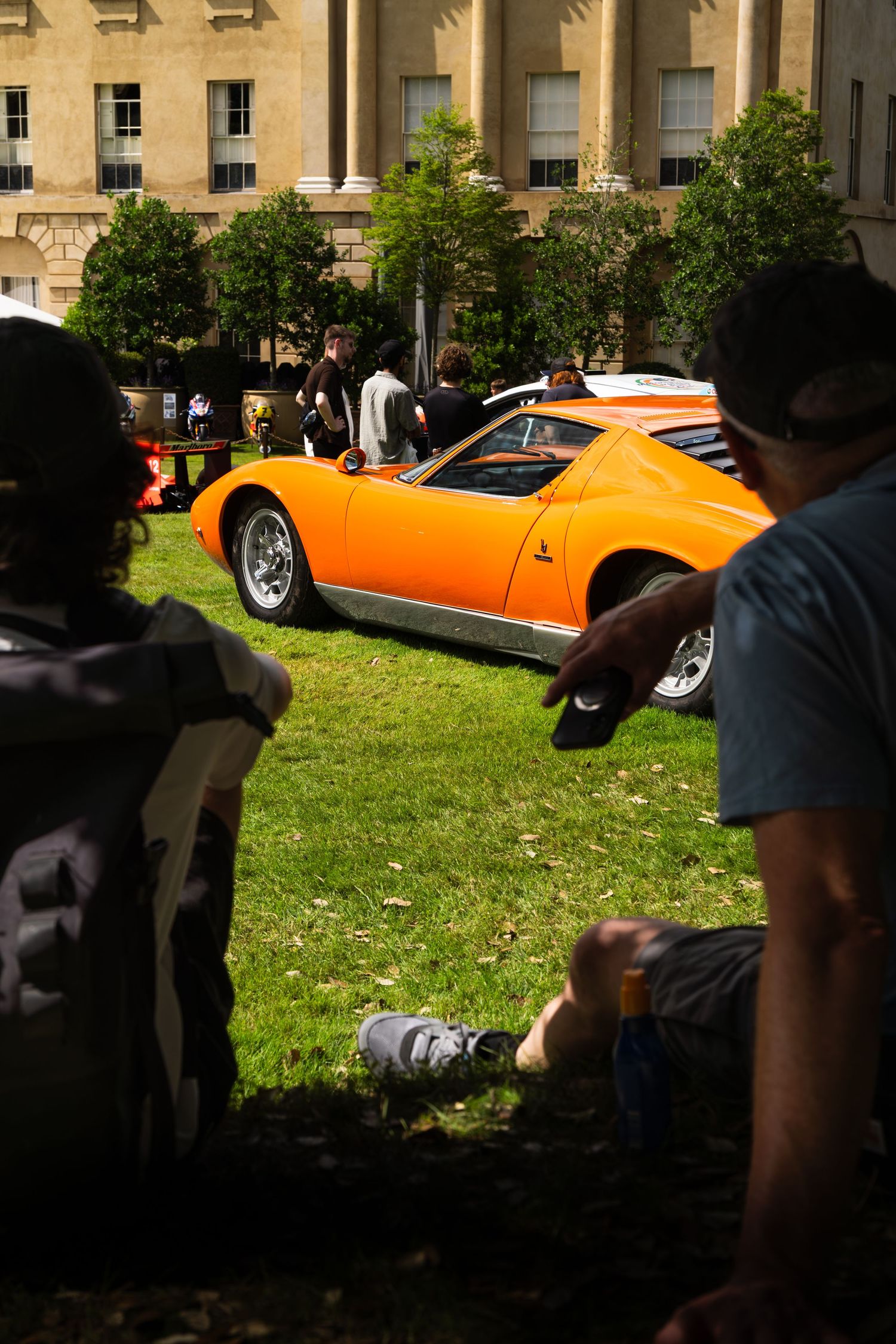 Spectators sit on grass admiring a classic orange Lamborghini Miura at an outdoor vintage car show near a historic building.
