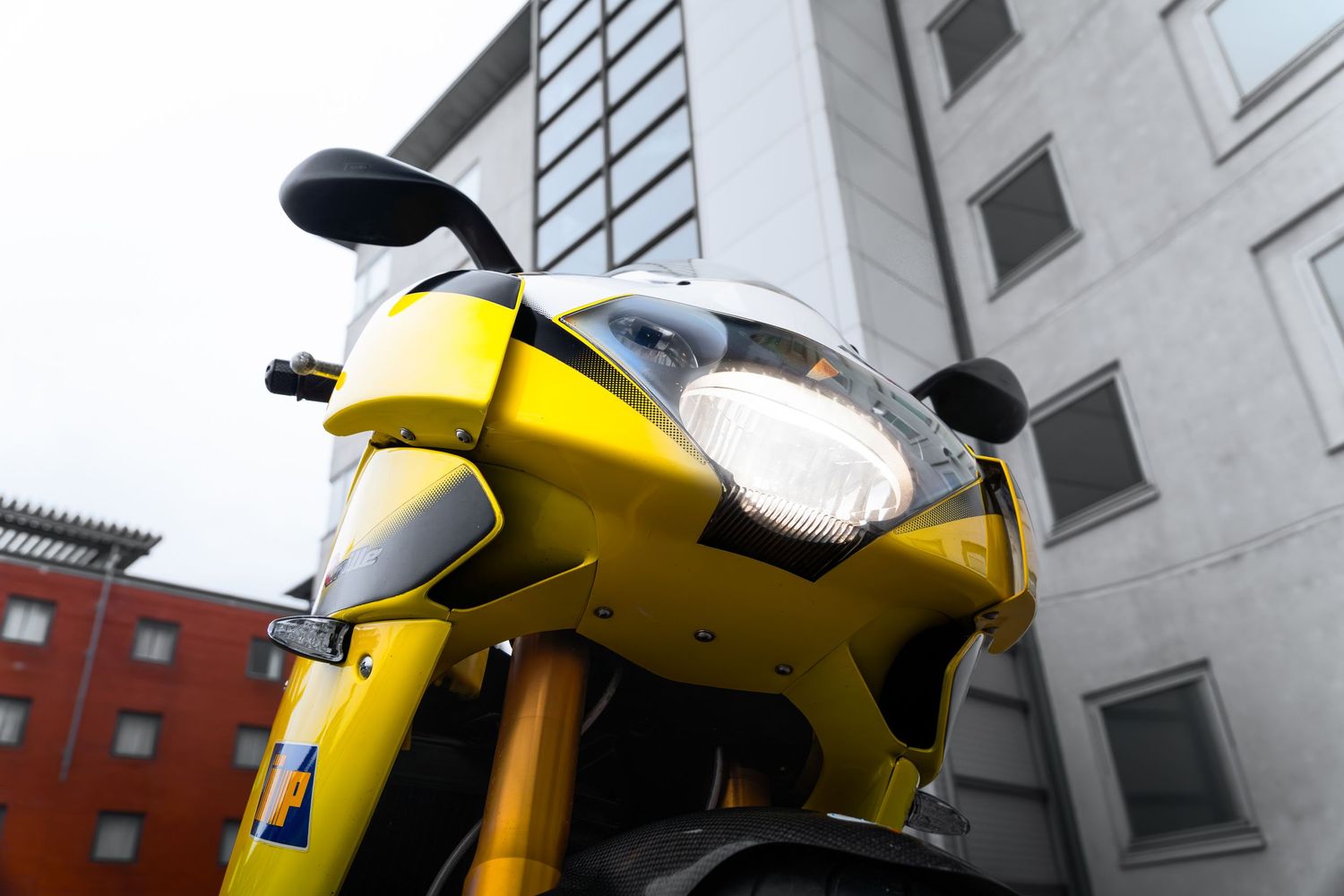 Low angle front view of yellow Aprilia sport motorcycle headlights against grey building.
