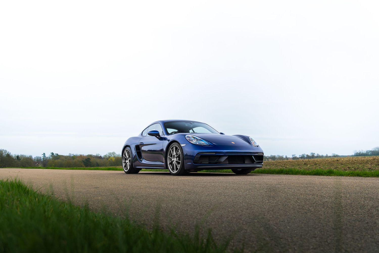 Blue Porsche 718 Cayman parked on a tarmac path surrounded by green countryside under grey sky.