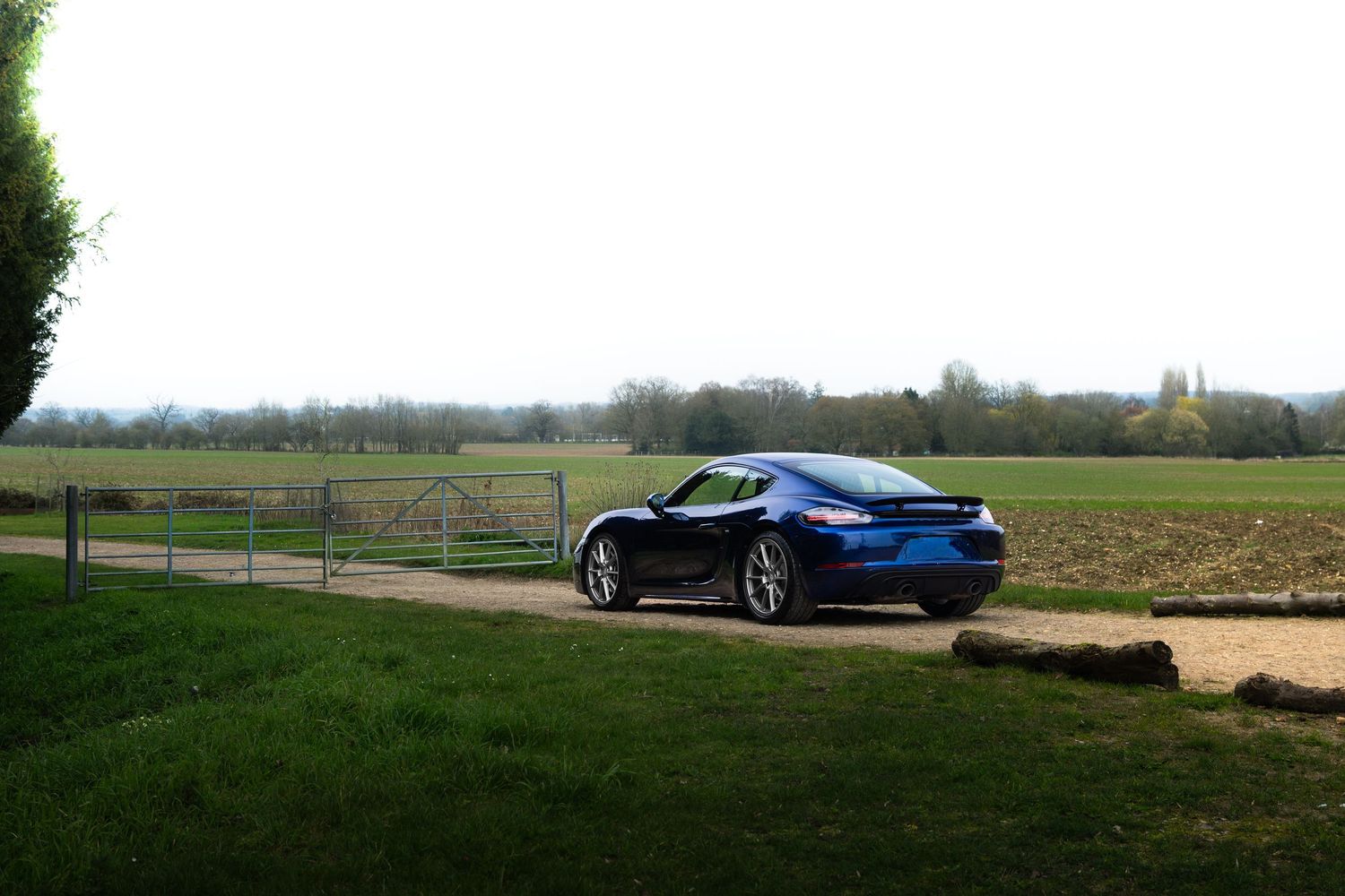 Blue Porsche 718 Cayman parked near a farm gate with open countryside and clear sky behind.