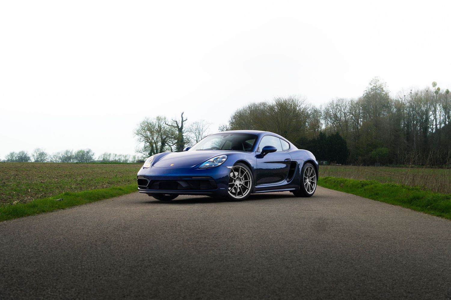 Blue Porsche 718 Cayman centered on a country road with green grass and trees behind.