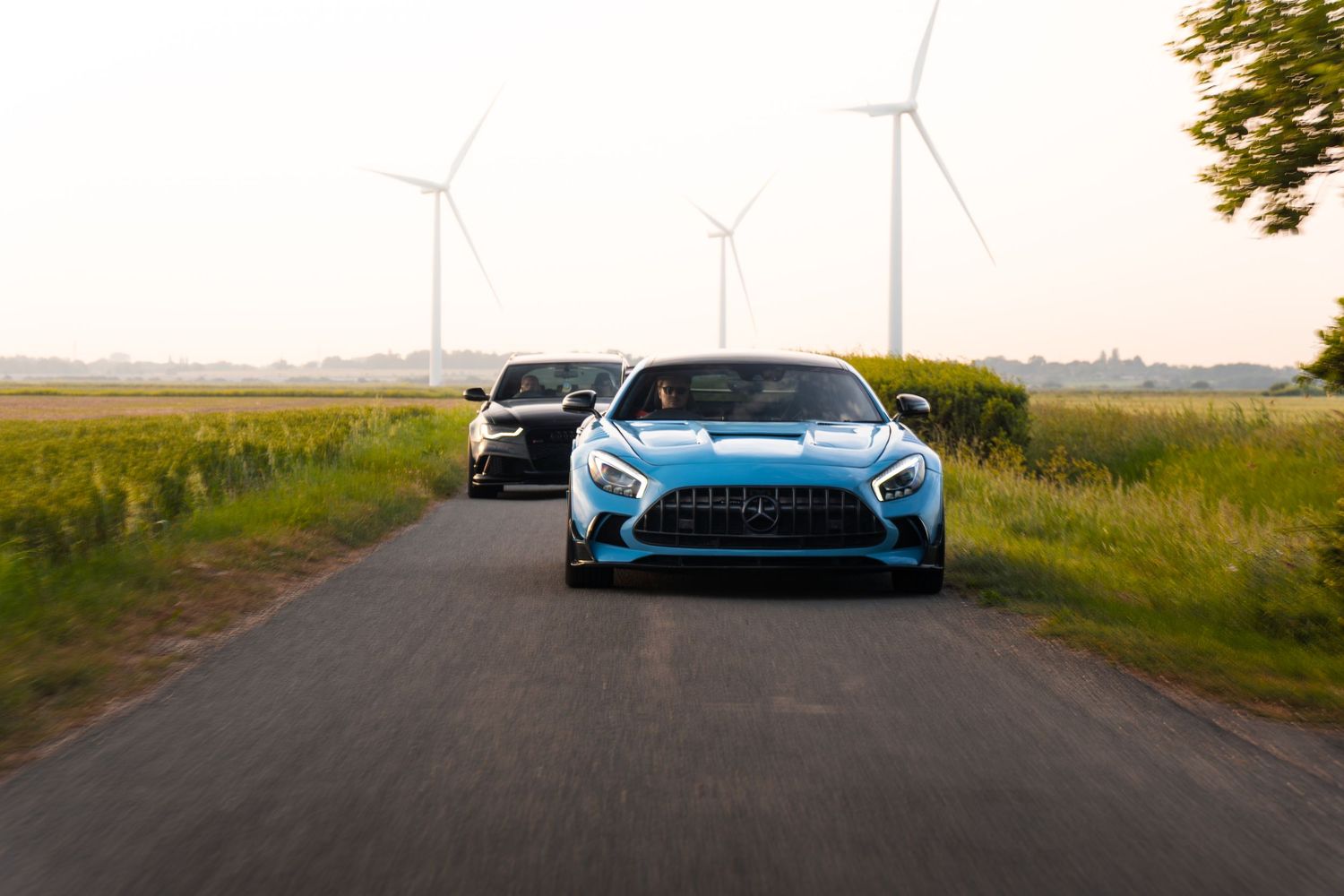 Blue and black Mercedes-AMG GT sports cars driving on a rural road with wind turbines.