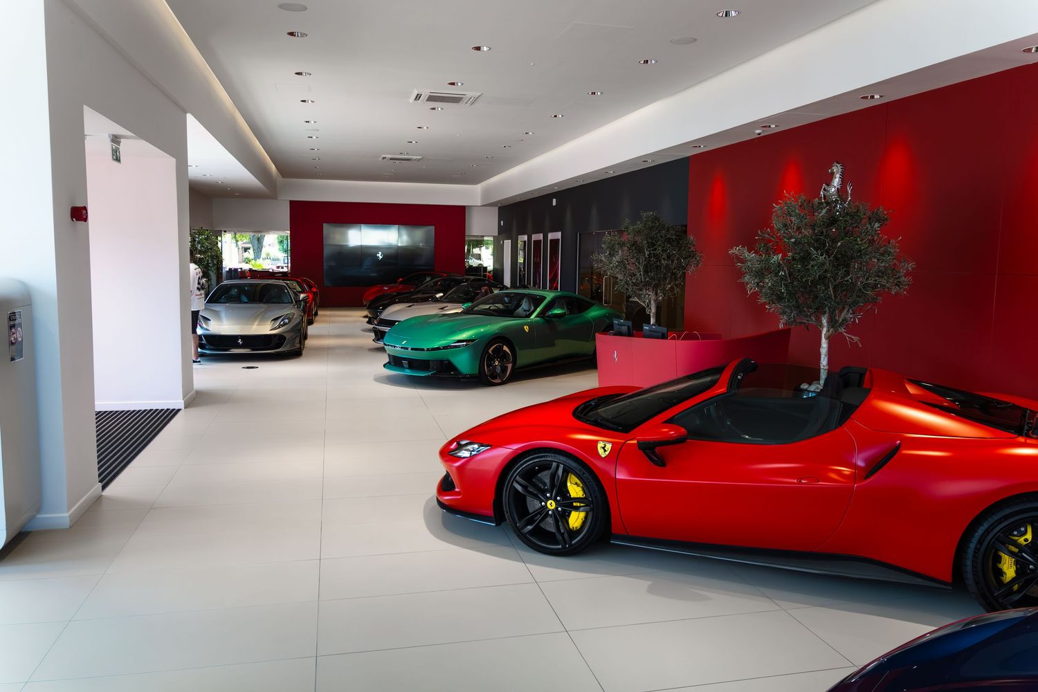 Luxury Ferrari showroom displaying red, green, and gray sports cars on white tile floors with red accent walls and indoor trees.