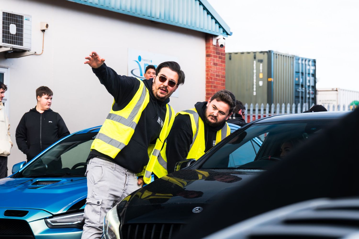 Two men in yellow hi-vis vests directing cars at an outdoor automotive meet event.