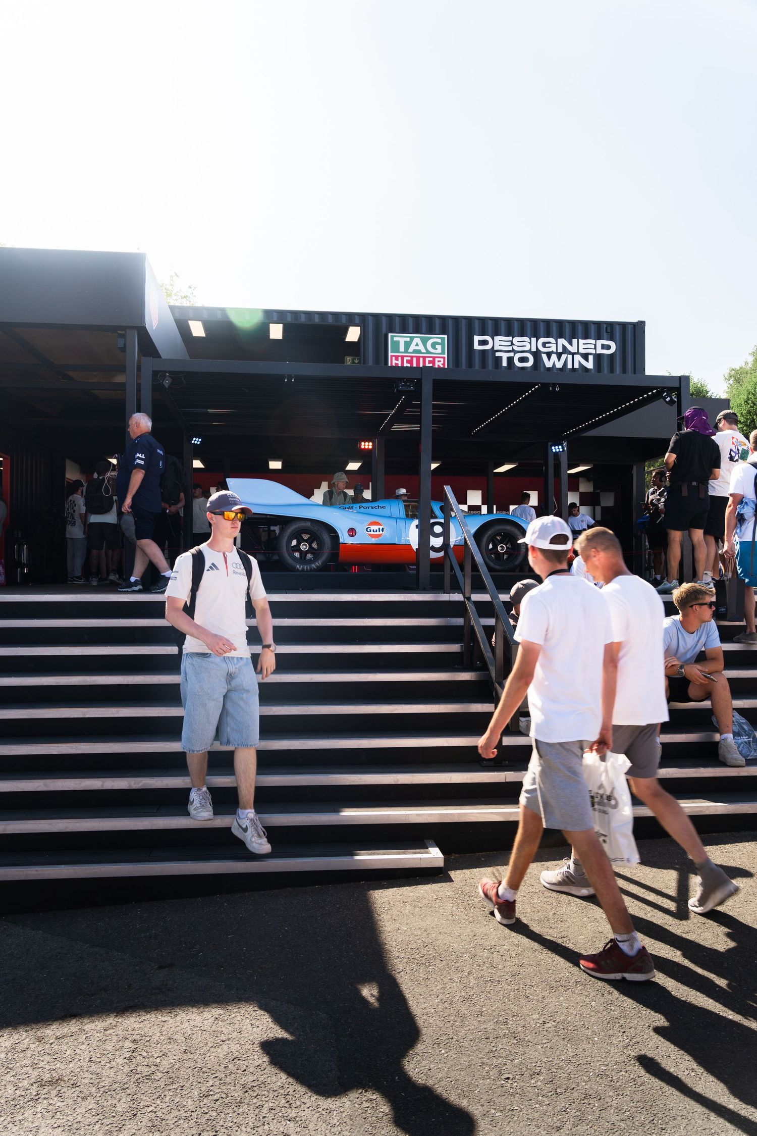 Visitors explore the TAG Heuer Designed to Win exhibit at a motorsport event, with a Gulf-liveried race car on display.