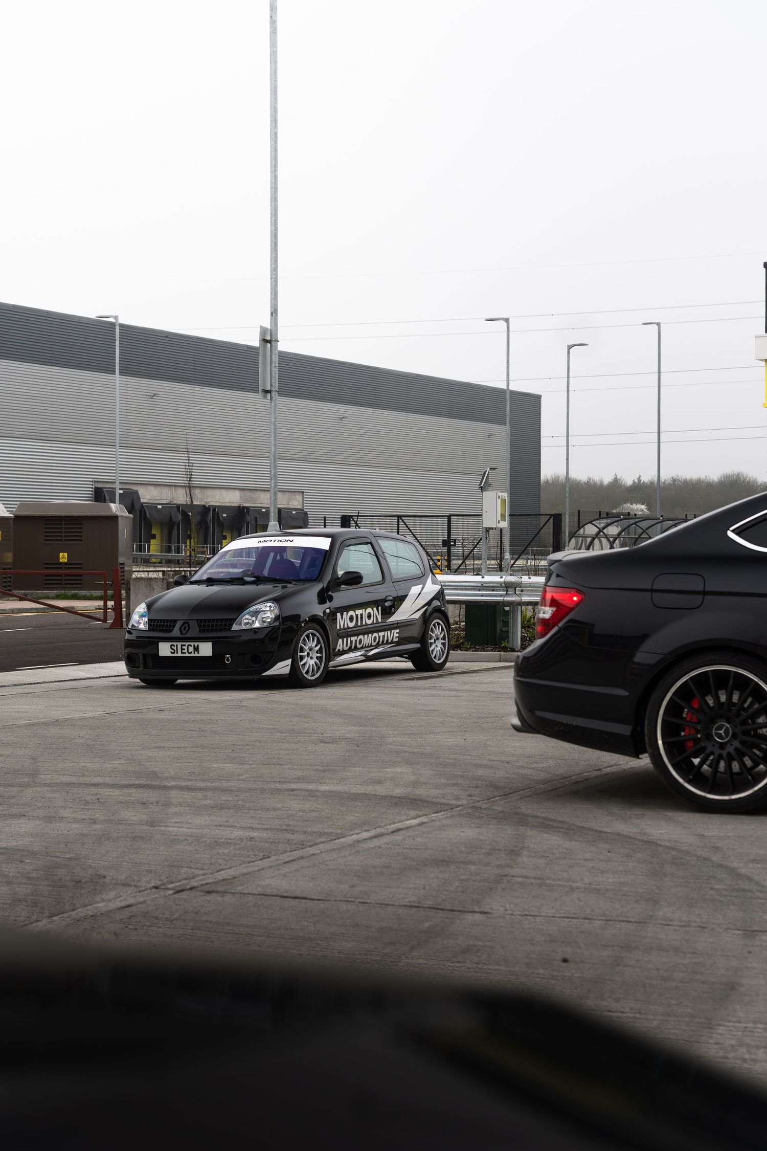 Black Motion Automotive branded Renault Clio parked in an industrial estate next to a dark Mercedes.