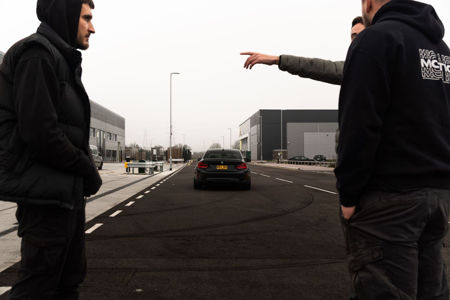 Two men in black jackets standing outdoors pointing at a dark car driving away in an industrial estate.