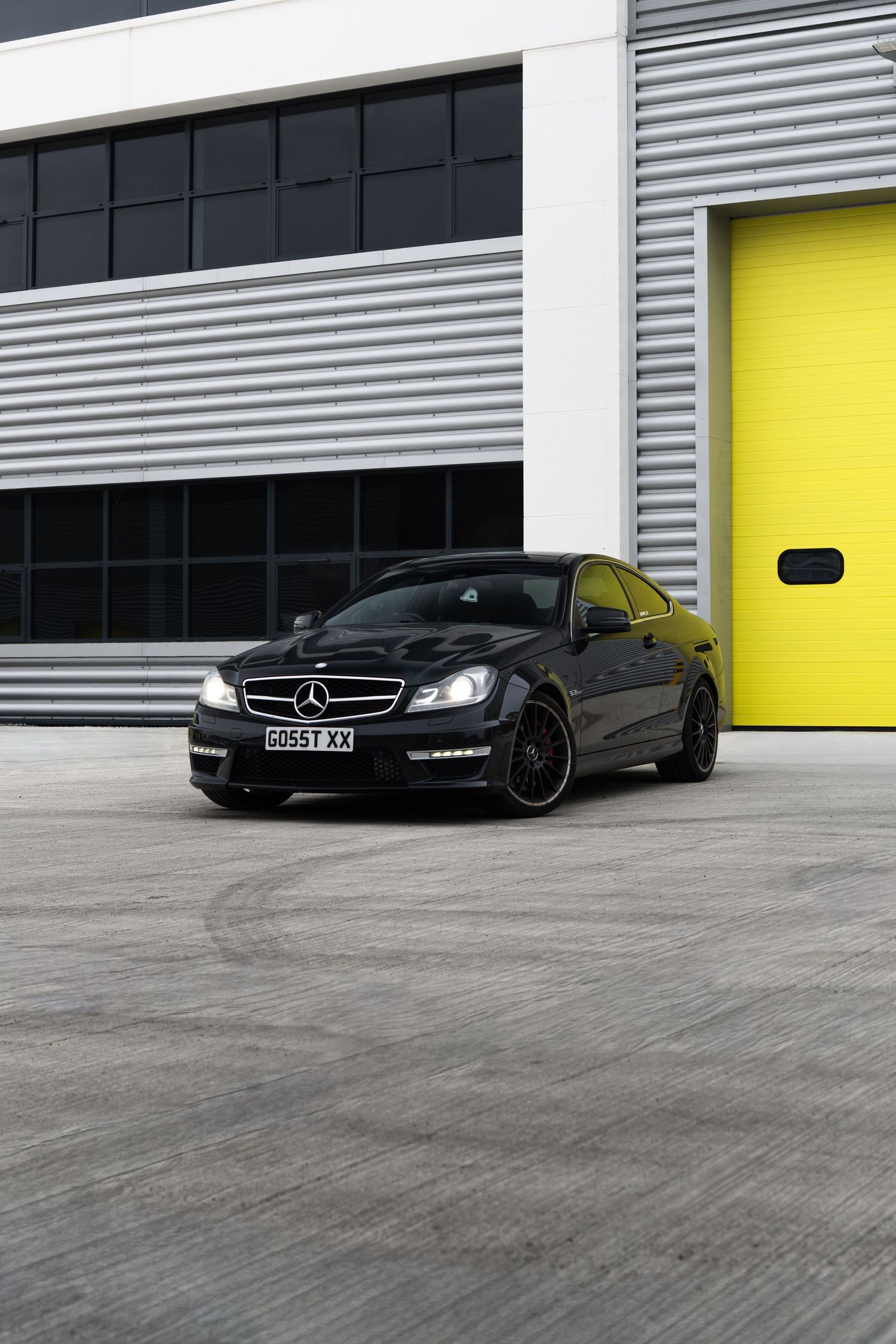 Black Mercedes-AMG C63 coupe parked on concrete apron beside yellow roller shutter garage door.