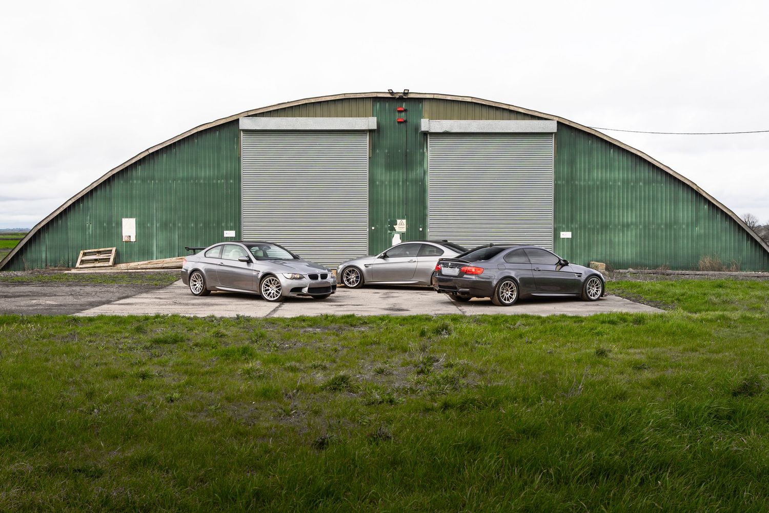 Three BMW M3 sports cars parked side by side in front of a large green arched hangar.