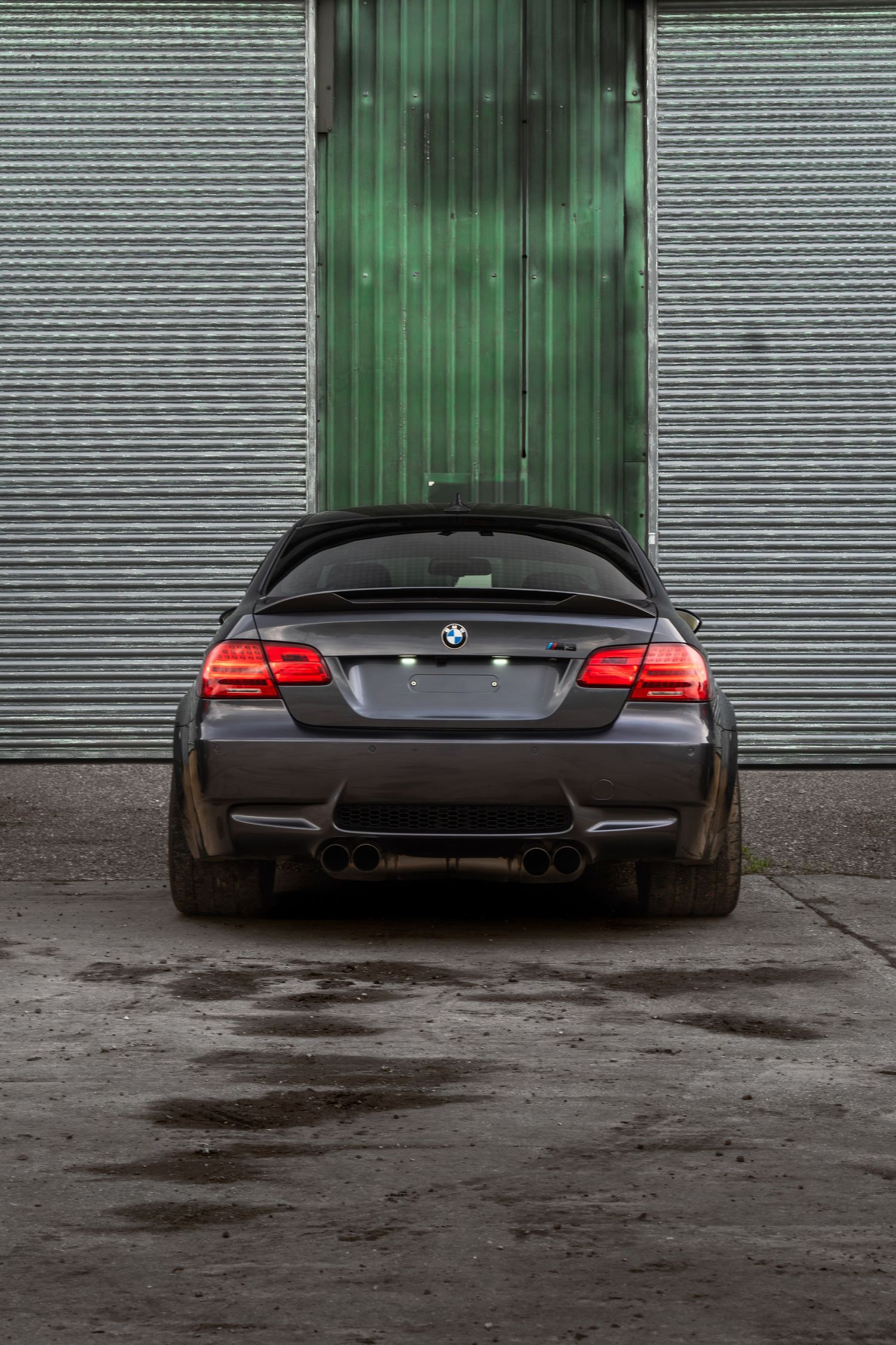 Rear view of a dark grey BMW M3 coupe with quad exhausts parked by green garage doors.