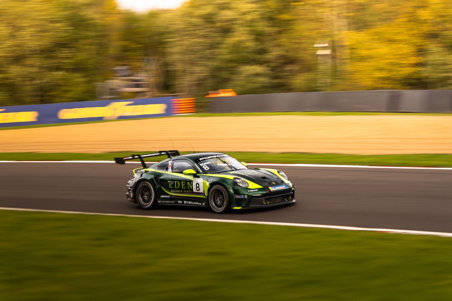 Green racing car number 8 speeding on a track with autumn trees in the background, captured with motion blur.