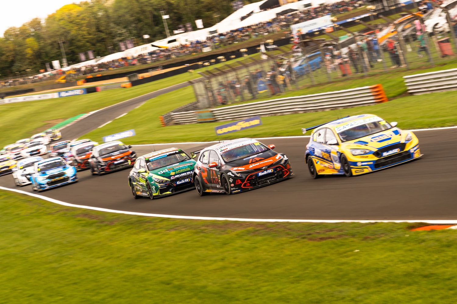 Multiple touring cars race through a sweeping corner at a packed British Touring Car Championship circuit.