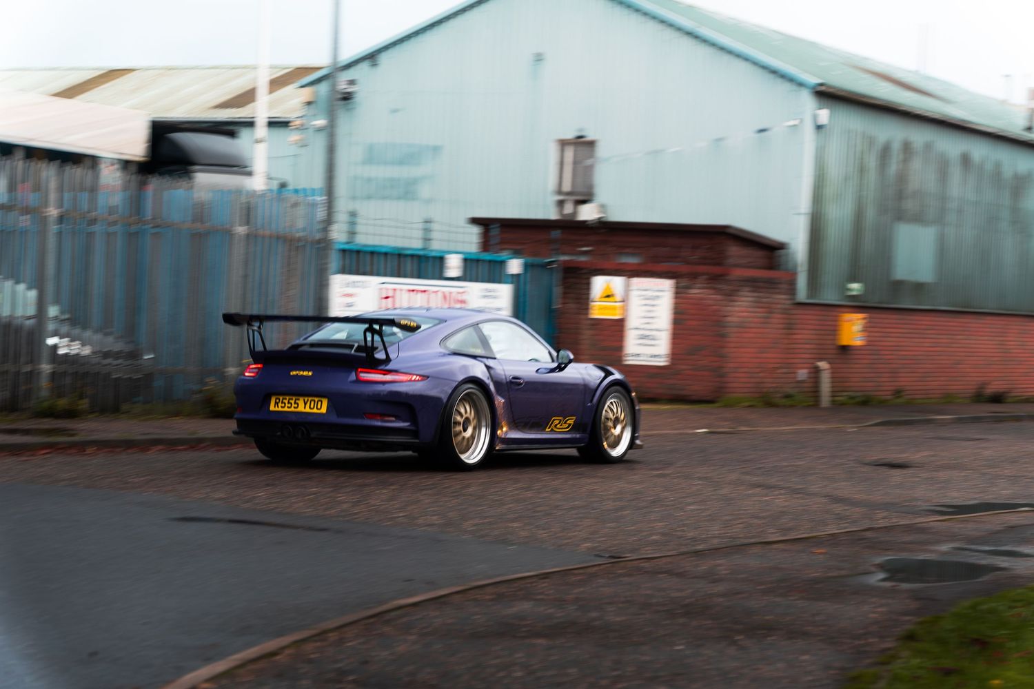 A purple wide-body Porsche 911 GT2 with gold wheels driving past industrial units.