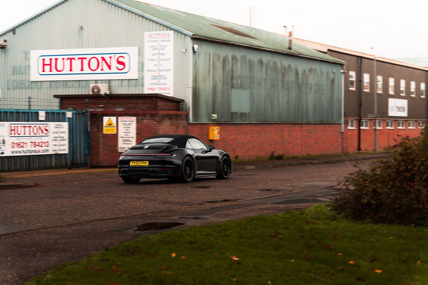 Black Porsche 911 parked outside Huttons industrial building in the UK on a cloudy day.