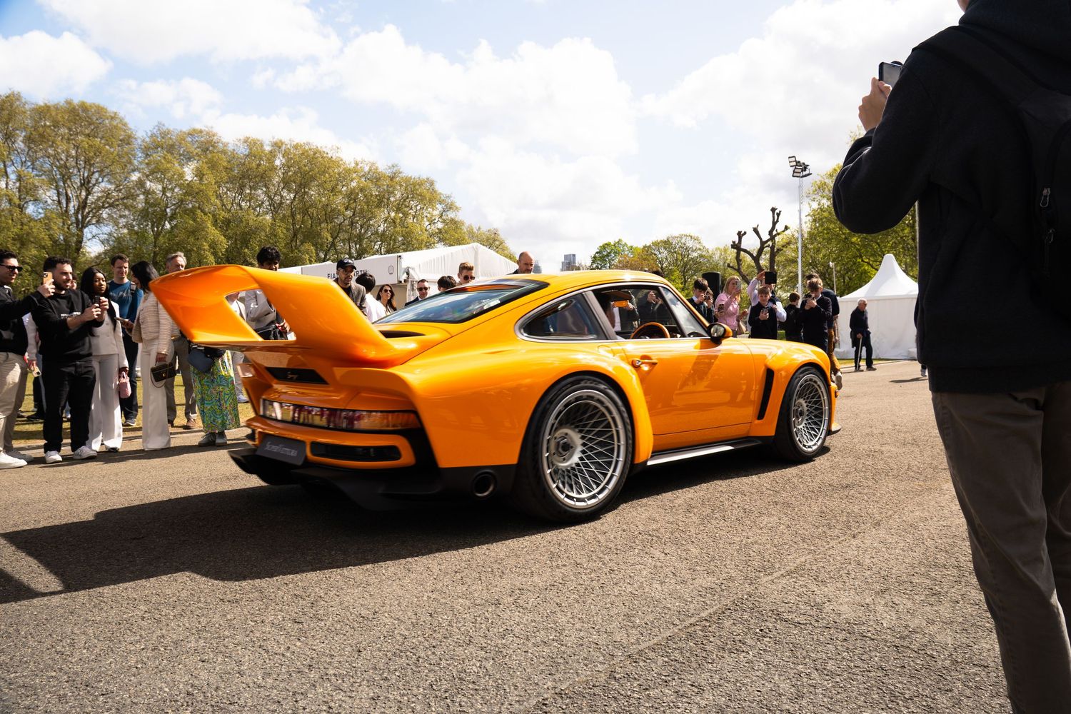 Rear view of orange RWB Porsche 930 Turbo surrounded by spectators at outdoor car event.