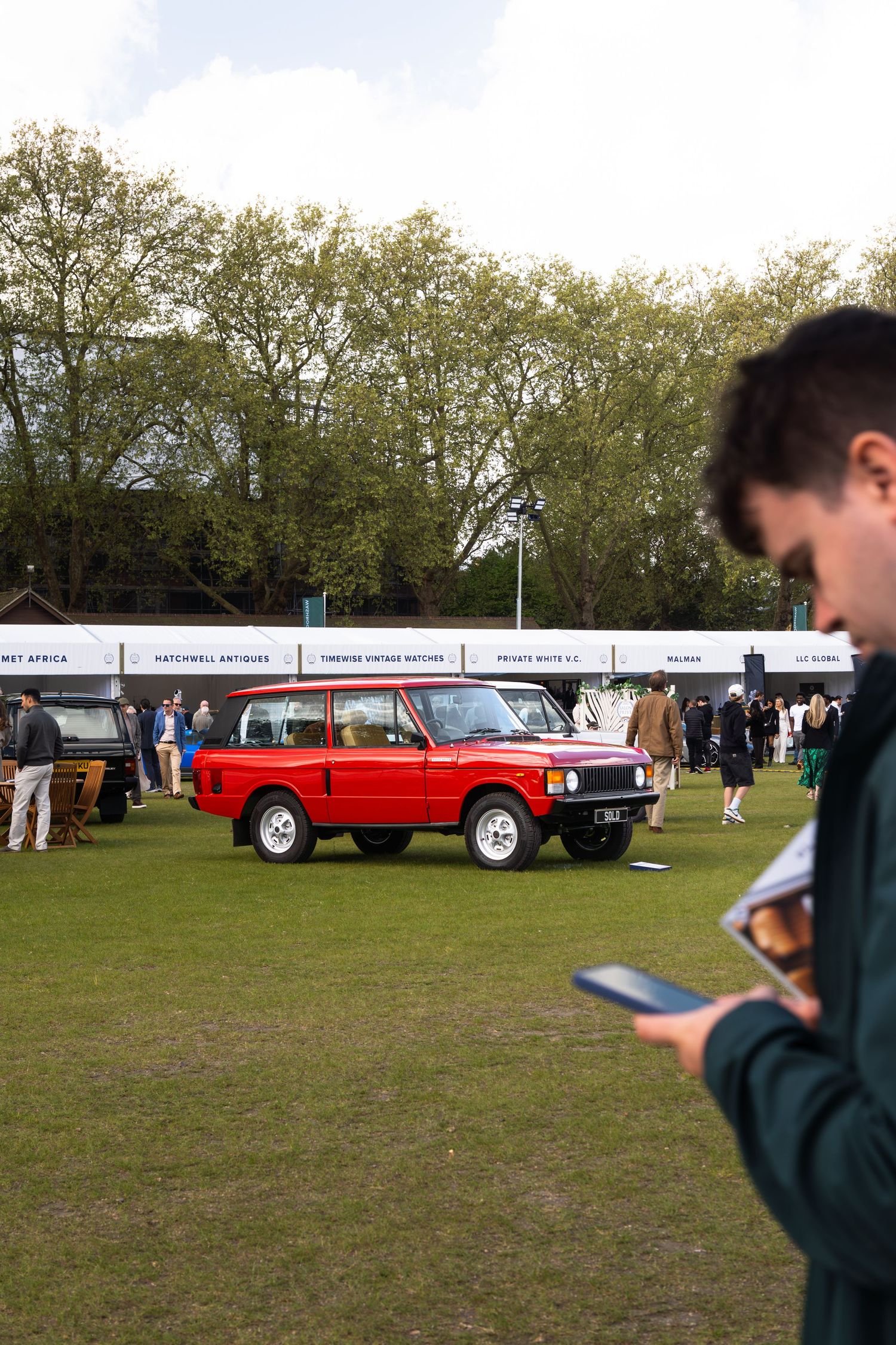 Classic red Range Rover on display at an outdoor vintage car show with spectators.
