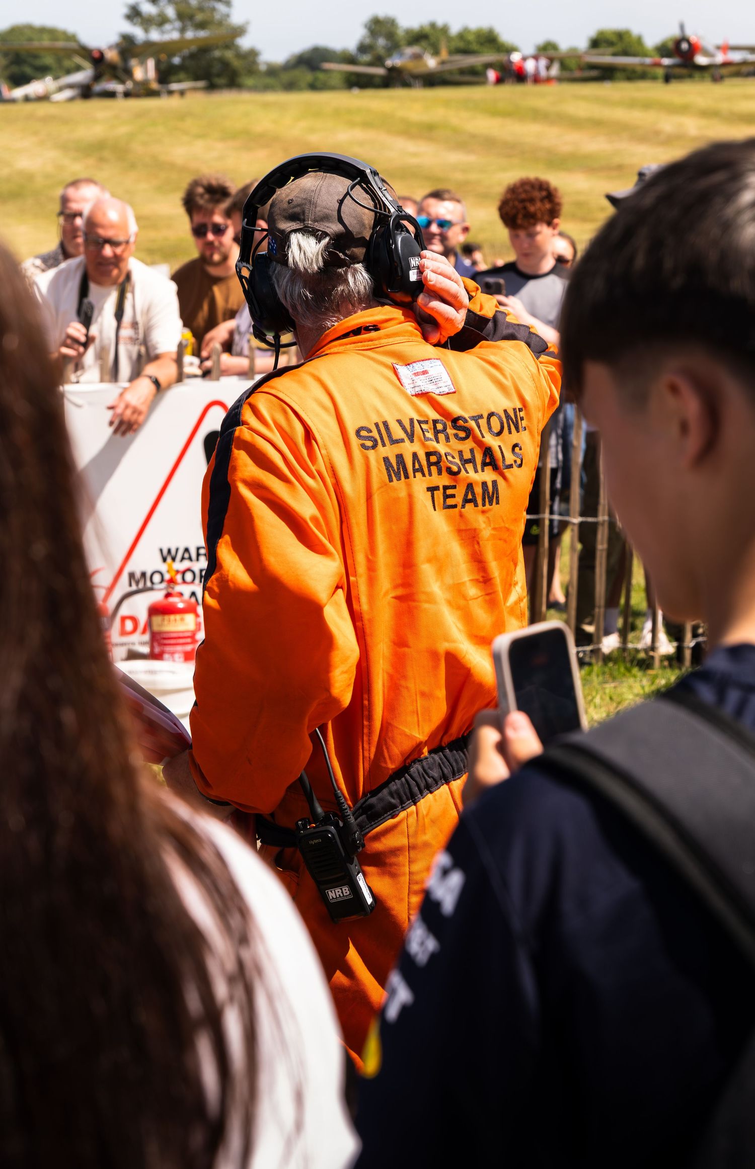 A Silverstone Marshals Team member in orange jumpsuit and headset stands among spectators at an airshow.