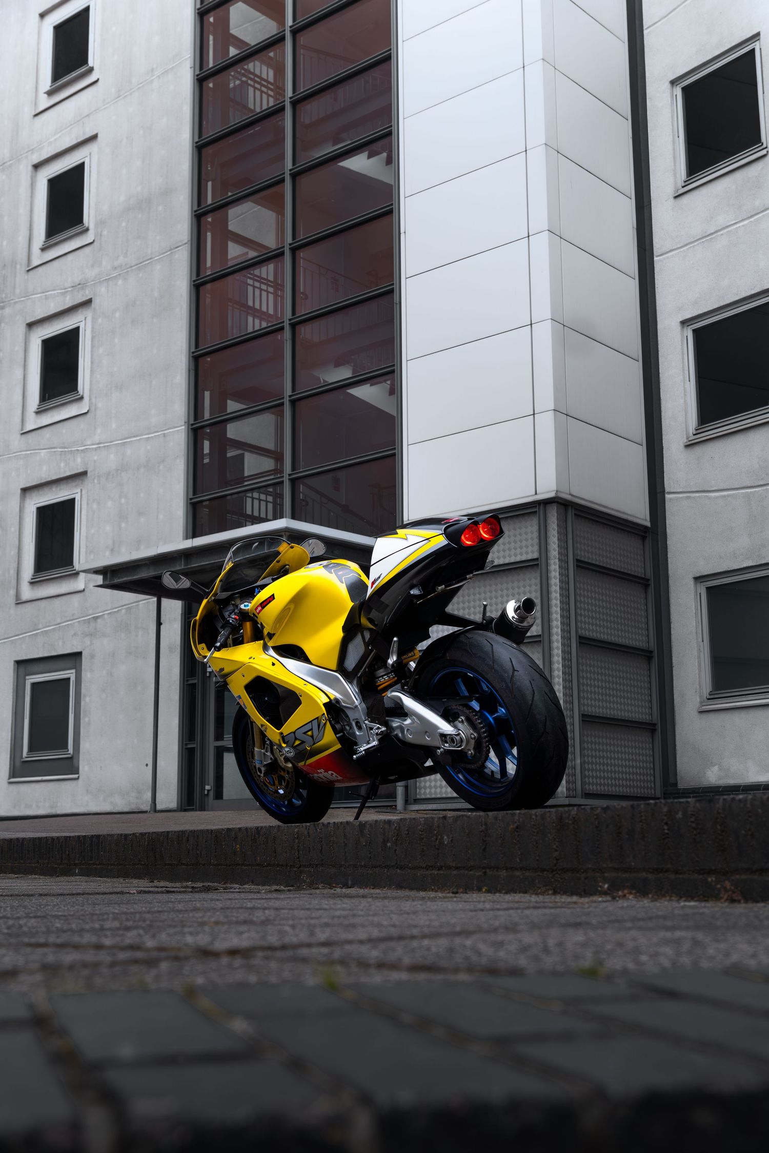 Rear view of a yellow Aprilia RSV1000 sportbike parked on cobblestones beside a modern building.