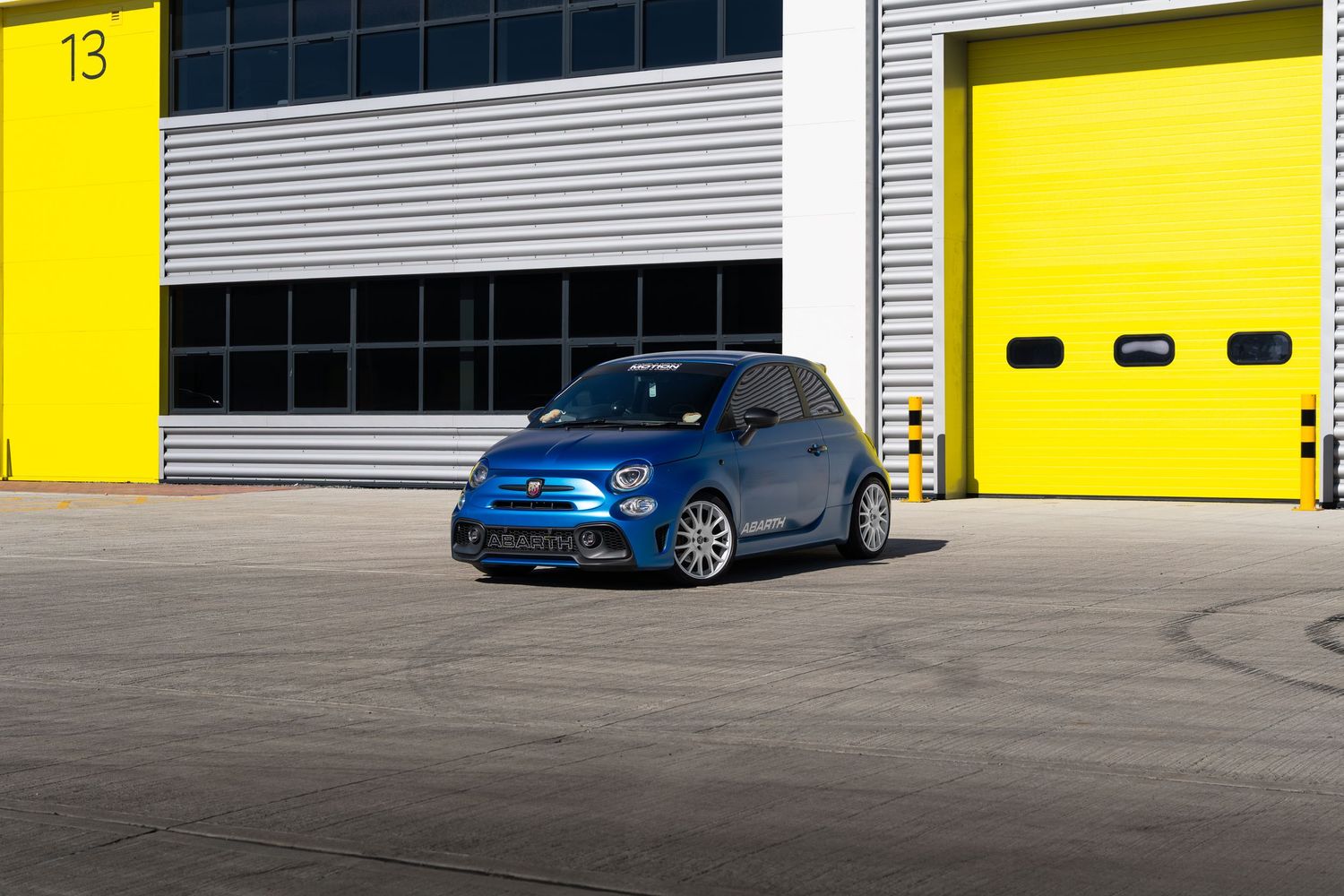 Blue Abarth 500 photographed outside numbered yellow garage doors at a racing circuit.