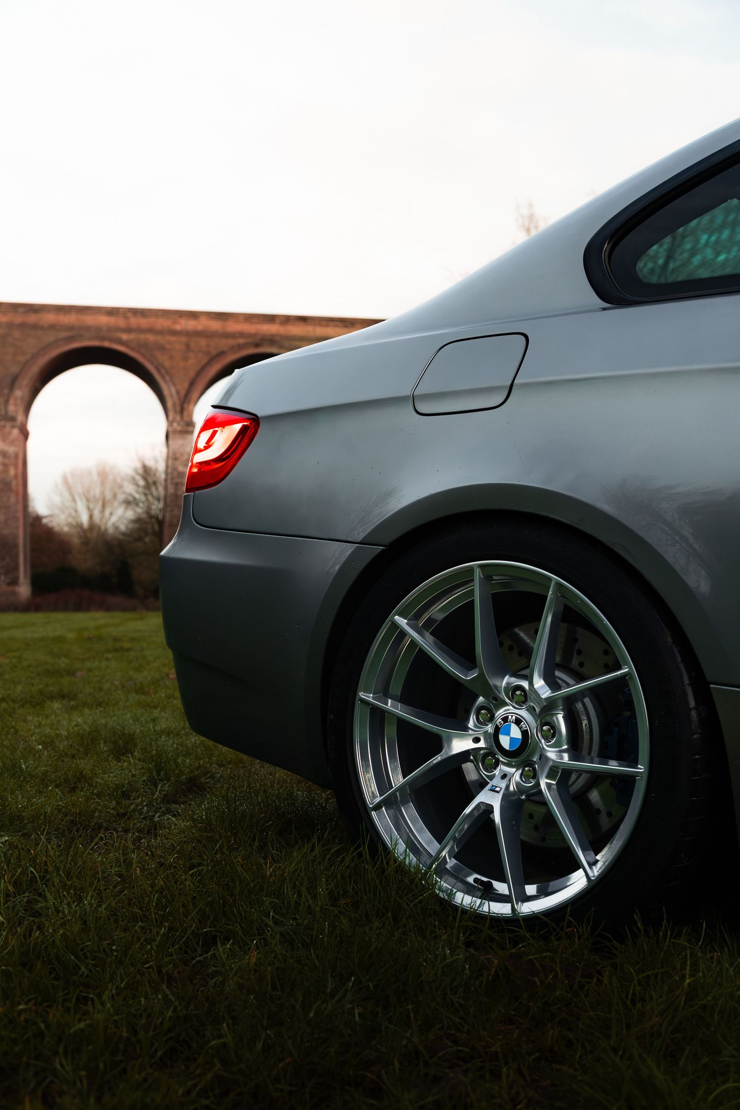 Close-up of BMW M3 rear quarter with chrome alloy wheel and red tail light near viaduct.