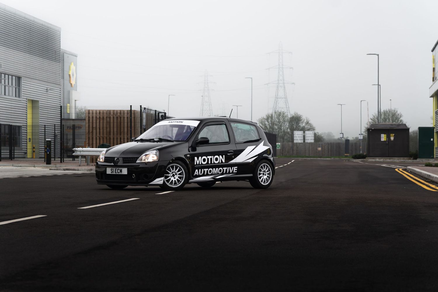 Motion Automotive branded black Renault Clio parked on a misty road beside an industrial building on an overcast day.